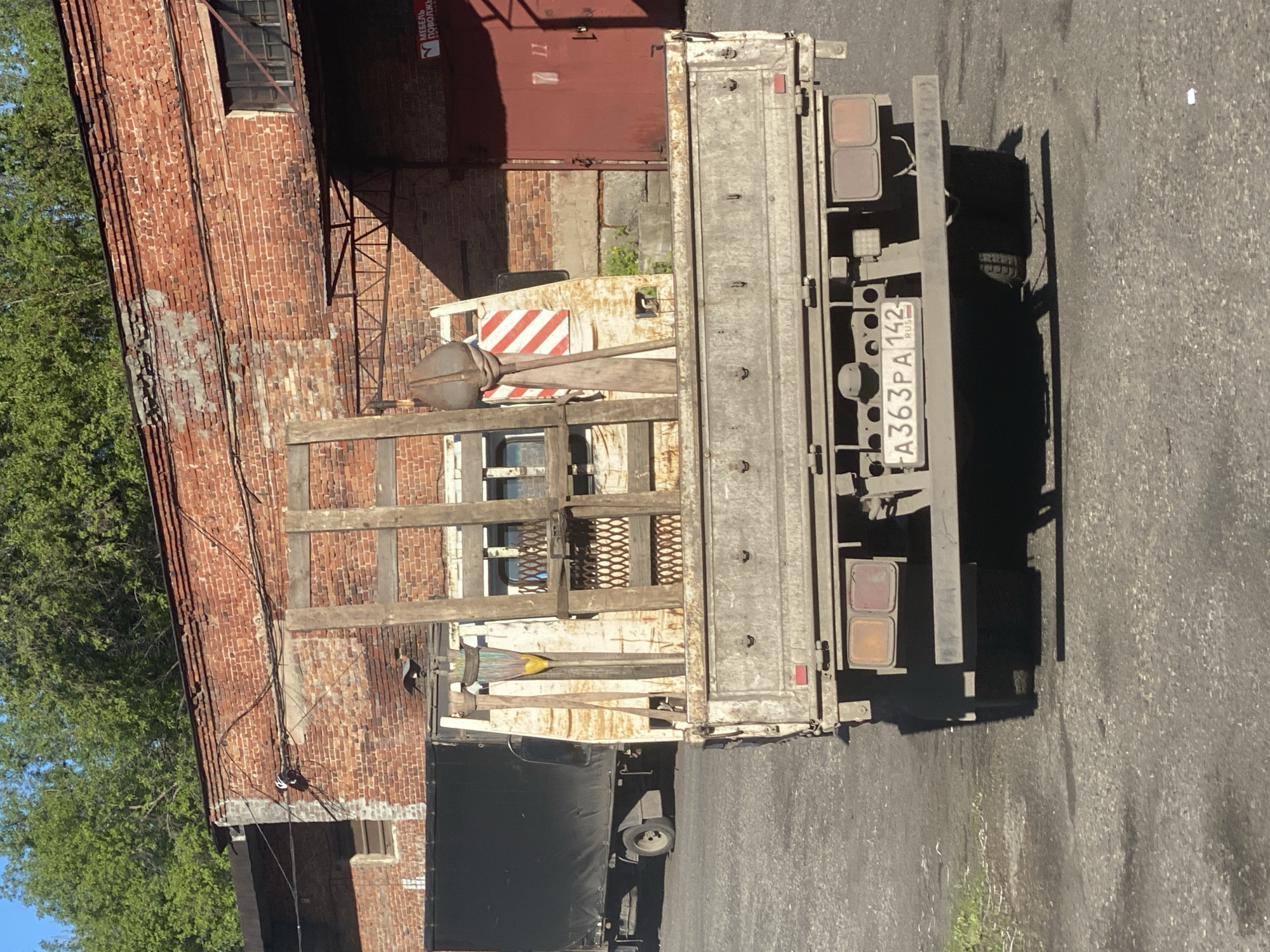 The image displays the rear view of a parked, well-worn, light-colored flatbed truck in what appears to be an industrial or commercial area of Novokuznetsk, Russia. The truck's bed is made of wood and metal, with various items including a large, spherical metal object on a stand, and a red-and-white striped barrier. Part of another vehicle, covered with a black tarp, is visible in the lower foreground.
The truck is positioned next to a tall, weathered brick building, likely an old factory or warehouse, with several framed windows and a large red metal door or shutter. Electrical cables are strung along the building's brick exterior. On the red door/shutter, a small red sign is affixed, showing partially obscured Cyrillic text: "МЕБЕЛЬ" (Furniture), "ПРОВОЛКА" (Wire), and "ВОЛОКН..." (likely "ВОЛОКНА" - fibers).
The scene is illuminated by bright daylight, casting strong shadows on the asphalt ground, suggesting it is mid-day or early afternoon on a clear, sunny day. Green foliage from trees is visible to the left of the brick building.
The truck's Russian license plate, "A363PA 142 RUS", is clearly visible. The "142" region code corresponds to Kemerovo Oblast, where Novokuznetsk is located. No people are visible in the image, contributing to a utilitarian and somewhat quiet atmosphere.