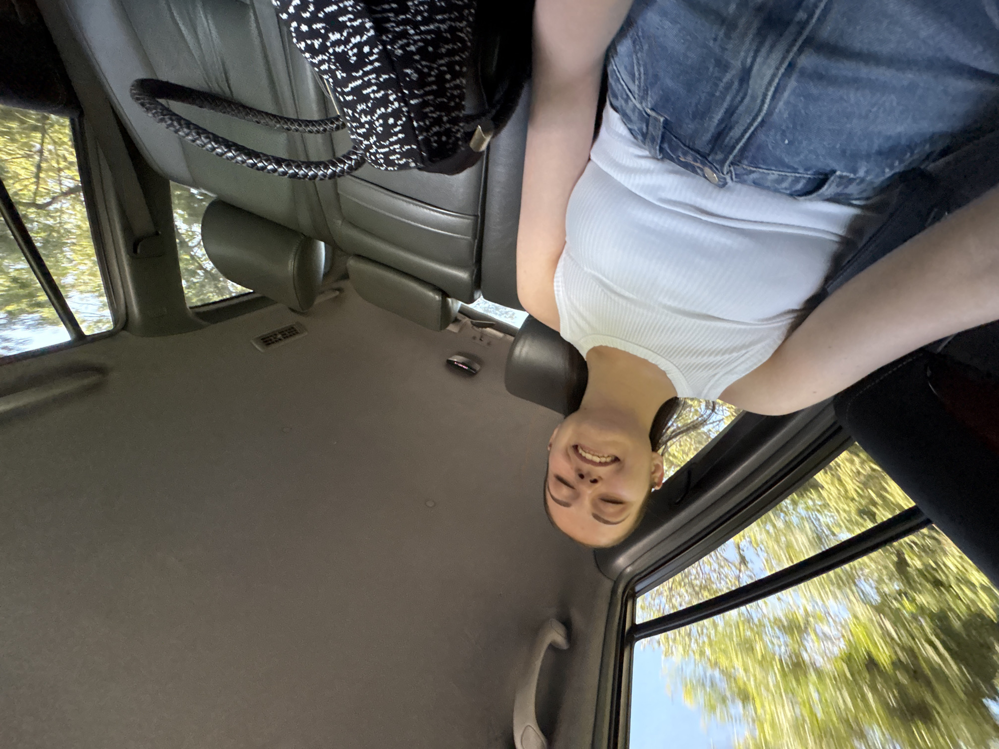 A young woman is captured in a playful, upside-down pose inside a moving vehicle, with her head oriented towards the bottom of the frame. She has a cheerful, wide smile, and her eyes are squinted in amusement. She is wearing a white ribbed tank top and blue denim jeans.
The setting is the interior of a car, likely an SUV or similar vehicle, characterized by dark grey, possibly leatherette, seats and a light grey fabric ceiling. A black and white patterned backpack with a distinctive braided strap rests on the seat beside her. Other interior details include a light-colored grab handle near a window, a small metallic light fixture on the ceiling, and a dark headrest. A small white rectangular sticker with illegible text is also visible on an interior panel.
Through the vehicle's windows, the bright daylight illuminates a scene of blurred green foliage, indicative of trees, against a clear blue sky. The blurring suggests the vehicle is in motion. The bright lighting indicates it is daytime with sunny weather. The overall mood of the image is one of lightheartedness and joy, possibly during a relaxed journey in Burgas, Bulgaria.
