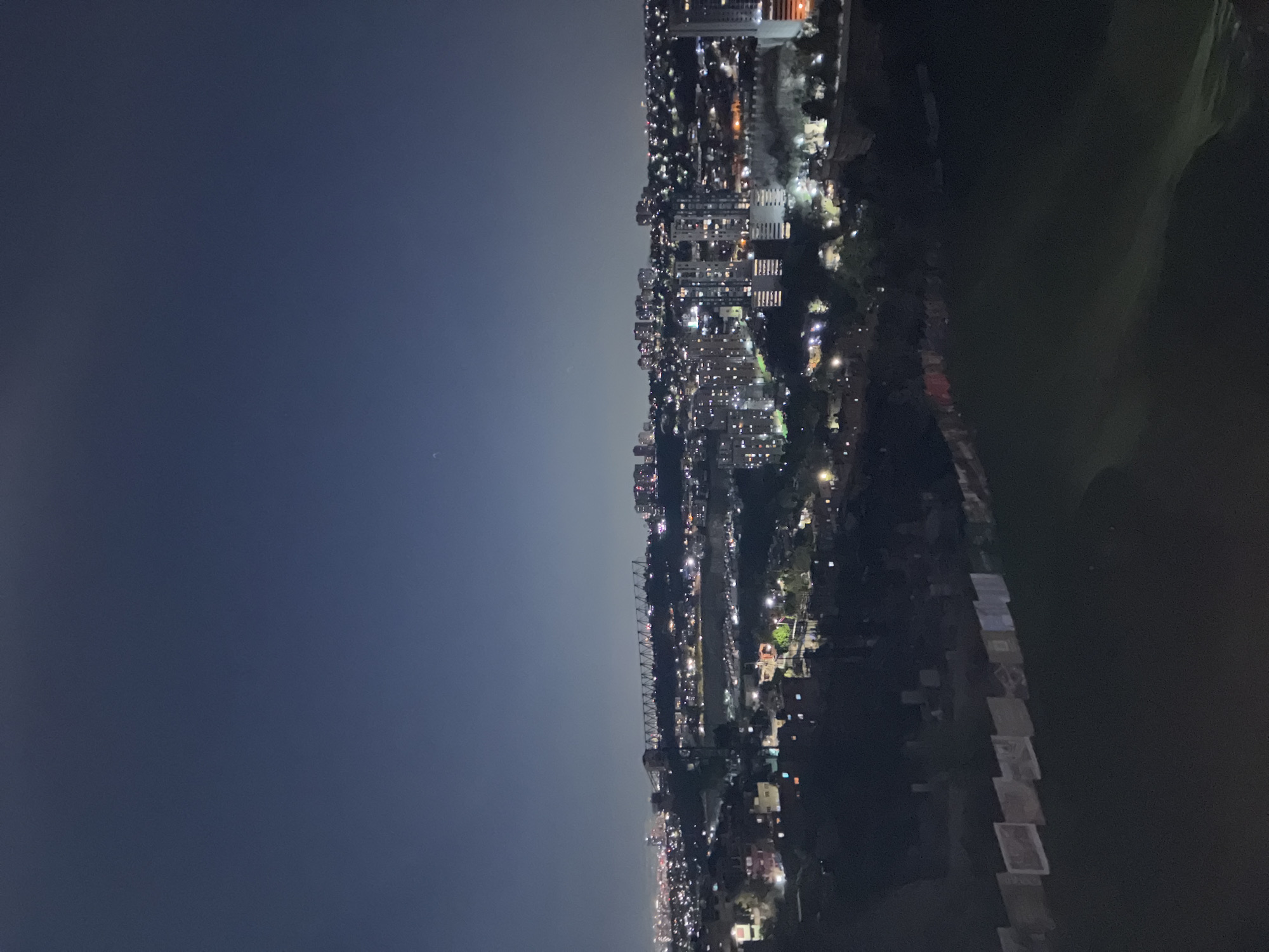 The image depicts a nighttime view of Huixquilucan de Degollado, Mexico. The left side of the frame is dominated by a dark blue sky. A cityscape stretches along the right side, illuminated by scattered lights from buildings and streets. To the far right, a dark, indistinct area suggests a body of water or open space.