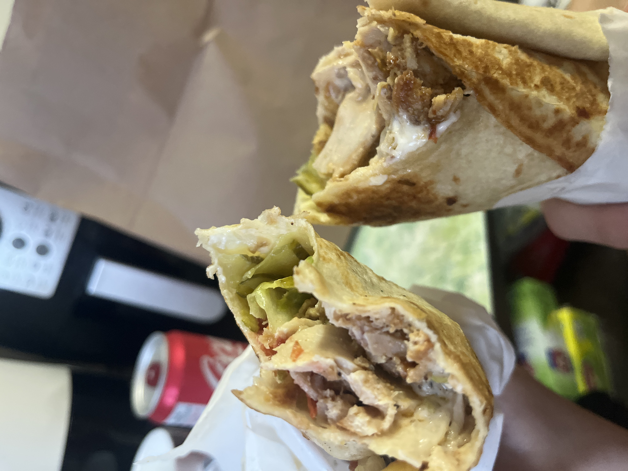 The image shows two shawarma wraps held in hands. One wrap has lettuce and pickles visible, along with chicken and sauce. A can of Coca-Cola is partially visible in the background near the bottom left of the frame. Other objects are present in the background as well. Location: Unknown, Unknown.