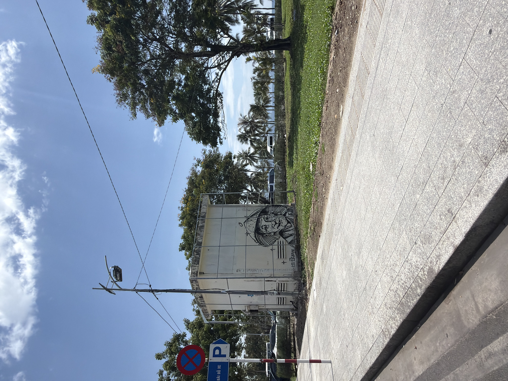 The image displays a sunlit outdoor scene, likely a street or promenade in Nha Trang, Vietnam. A gray paved sidewalk, composed of rectangular tiles, occupies the right side of the frame, bordering a strip of green grass and dirt on its left.
A large, light-colored utility box or small building stands prominently to the left of the sidewalk. Its visible side features a striking black and white graffiti portrait of an individual, possibly an older person with a hat or head covering, rendered in a graphic, detailed style. Below the portrait, on its right, there is some stylized, illegible text or a signature.
In front of the utility box, a utility pole rises, equipped with a street light fixture and several electrical wires that stretch across the clear blue sky. Attached to the pole is a sign with a blue square containing a white 'P' for parking, which is crossed out by a red circle with a diagonal line, indicating "No Parking." Below this, Vietnamese text reads "BÃI ĐỖ XE," which translates to "Parking Lot," confirming the local context. A red and white striped barrier pole is also visible near the sign.
Beyond the grass and utility box, a lush environment of trees is evident. A large leafy tree extends from the top left, and a dense line of palm trees stretches across the middle ground, partially obscuring what appears to be a body of water or coastline in the distance. The bright sunlight suggests it is daytime, with scattered white clouds in the sky. No people or specific activities are depicted.