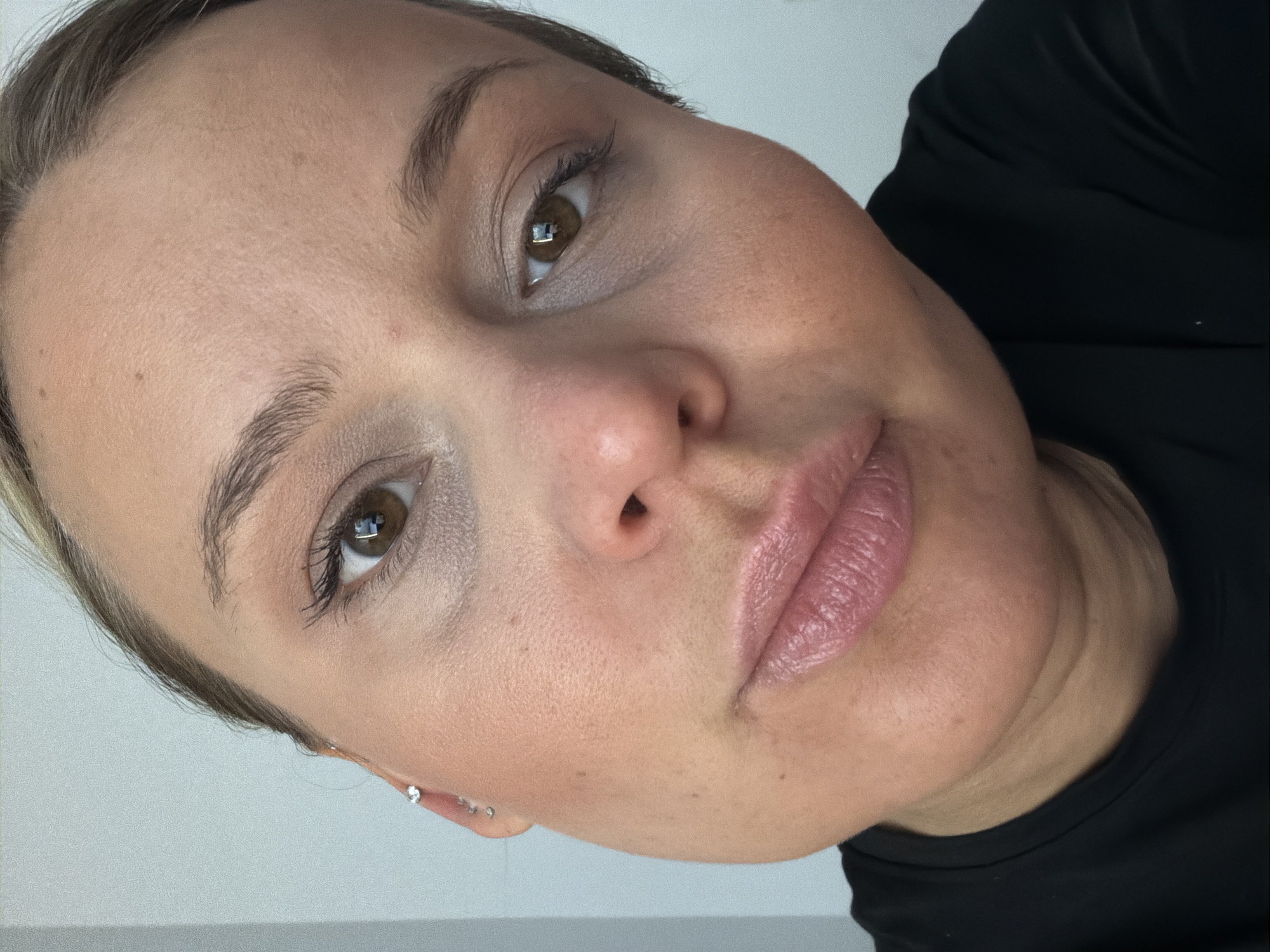 A close-up, angled portrait features a woman with fair skin, light brown eyes, and short light brown hair. She wears a black shirt and earrings, her lips are a pale pink, and she has makeup on her eyes. The background is out of focus, with a white wall visible. The location is unknown, and there is no information connecting the image to Sarapul, Russia.