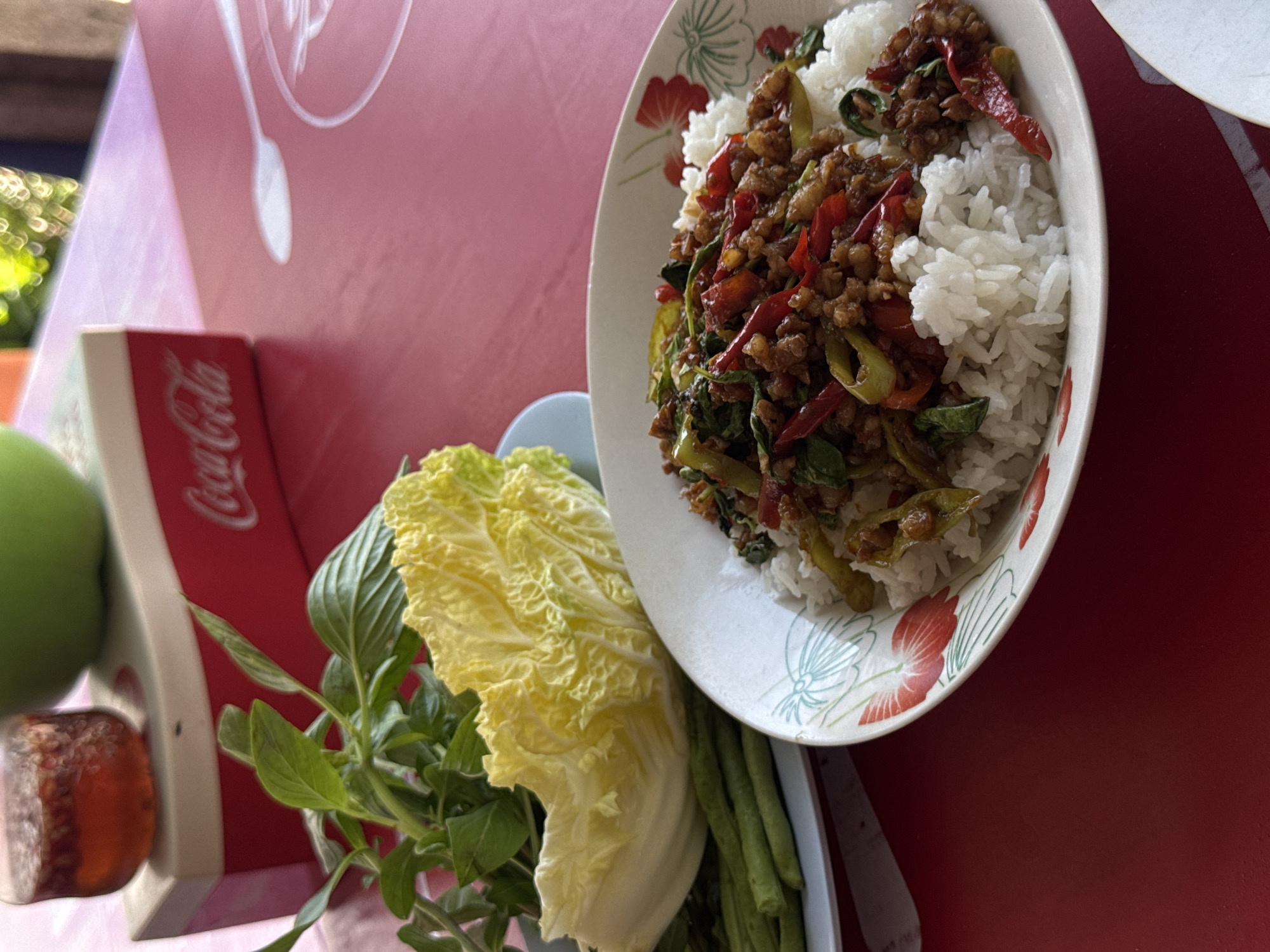 A vibrant Thai meal is presented on a bright red table, likely in Pattaya City, Thailand. The central focus is a white bowl with a red floral pattern, holding a serving of fluffy white rice topped with a generous portion of stir-fried Pad Kra Pao. This dish features minced meat, prominent red chili slices, green chilies or green beans, and fresh green basil leaves.
Adjacent to the main dish, a white rectangular tray holds an assortment of fresh vegetables intended as sides. These include a large, pale yellow-green leaf of cabbage (possibly Nappa cabbage), a bundle of aromatic green basil, and several long green beans.
On the far left, a red Coca-Cola box is visible, clearly displaying the "Coca-Cola" logo in white script. Beside the box, a round green fruit and a small reddish-brown item are also present. The scene is illuminated by natural daylight, suggesting an outdoor or semi-outdoor dining setting, possibly at a casual eatery or street food stall. No people are visible. The arrangement indicates a meal ready to be consumed.