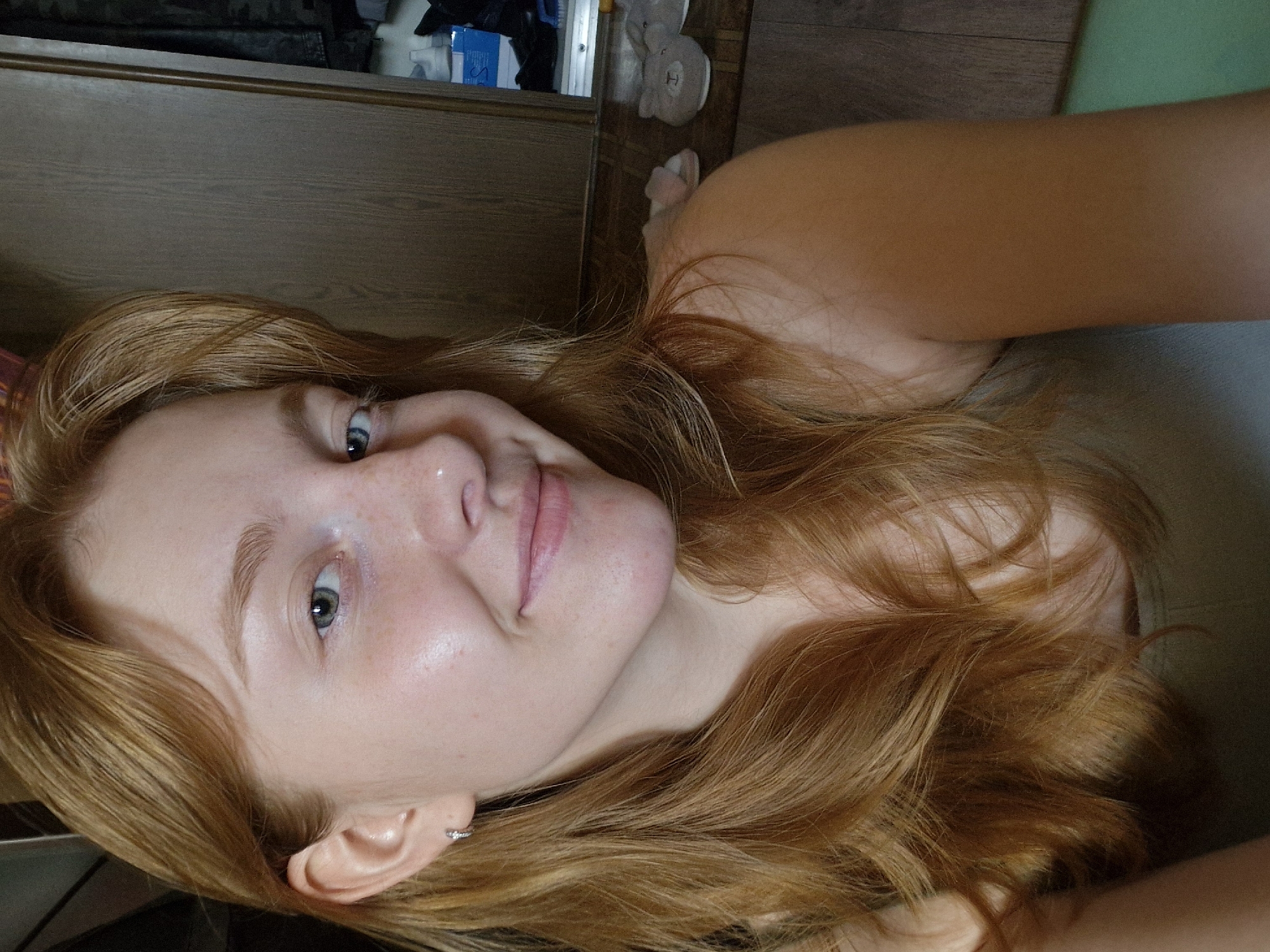 A young woman with auburn hair is taking a selfie in what appears to be a domestic environment. She is looking at the camera and smiling slightly. Her left ear, visible in the foreground, has a small hoop earring. Her skin is fair with visible freckles. She is wearing a light-colored tank top. Behind her, a wooden closet door is partially open, revealing some items inside. In the background, on a wooden surface, a pair of fuzzy slippers are visible. The lighting suggests an indoor setting, possibly a bedroom or bathroom. No specific landmarks are visible to pinpoint a location in Yekaterinburg, Russia.