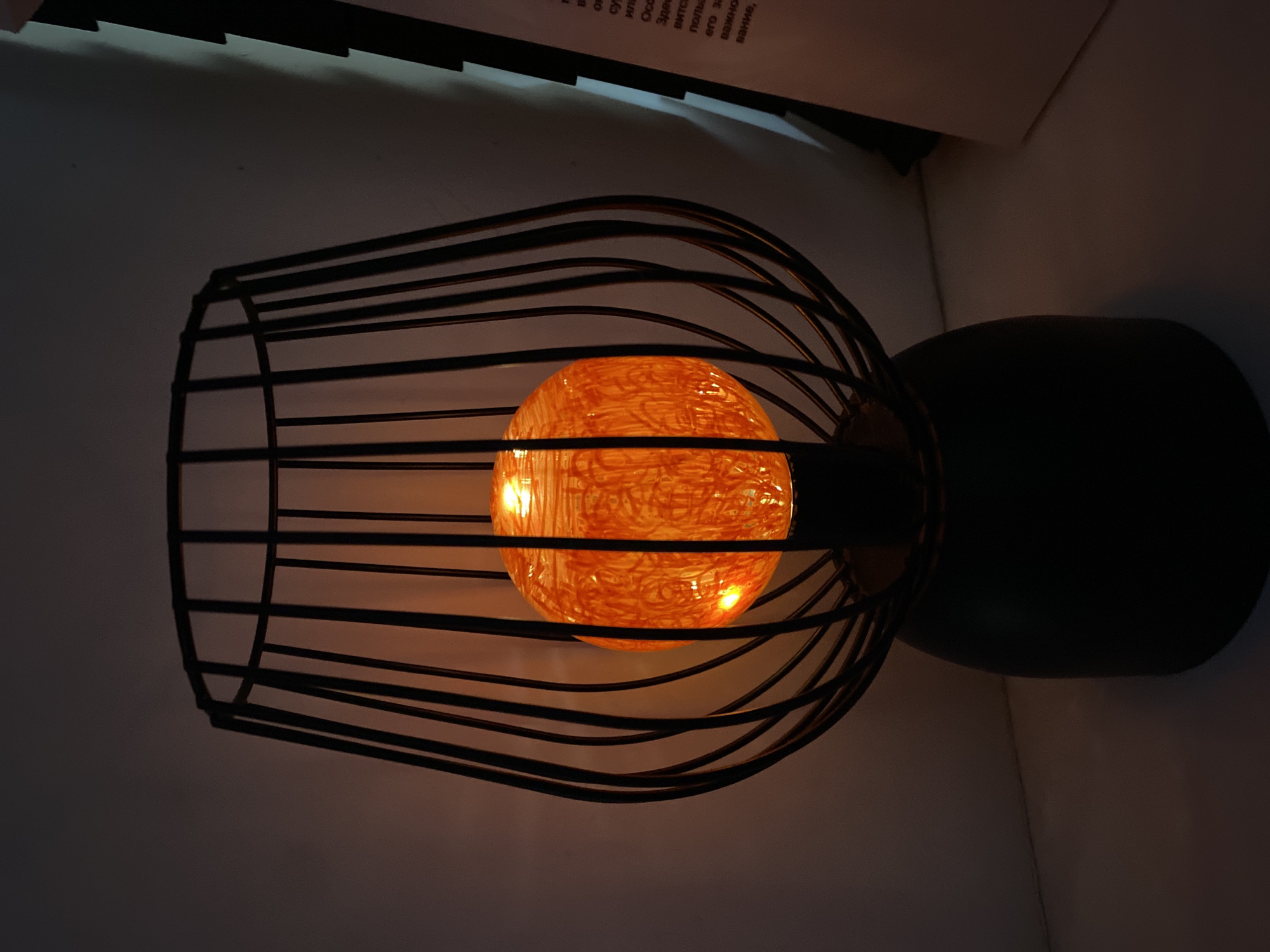 A black metal cage-like lamp encloses an illuminated orange spherical light. The lamp sits on a dark base. It is situated indoors. It may be located in Yekaterinburg, Russia.
