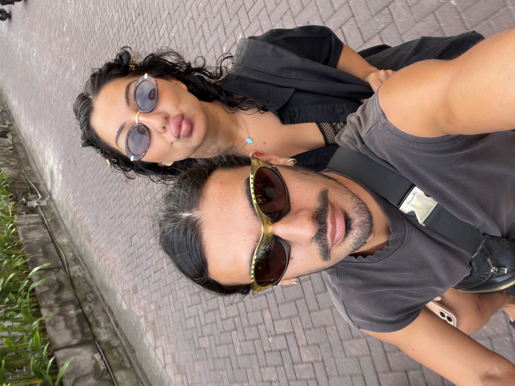 A selfie features two individuals, a man and a woman, taken outdoors during daytime in what appears to be North Kuta, Indonesia. The image is rotated, presenting the scene diagonally.
In the foreground, a man holds a light pink smartphone with dual cameras, suggesting he is taking the selfie. He has dark hair with visible grey strands, a beard, and a mustache. He wears distinctive olive-green framed sunglasses with dark lenses and a dark grey, possibly faded, t-shirt. A black strap with a silver buckle, likely from a bag or camera, diagonally crosses his chest.
Behind the man, a woman with dark, curly hair is visible. She wears round, tortoise-shell patterned sunglasses and has small gold-colored clips in her hair. Her lips are pursed in a 'duck face' pose, with a hint of her tongue visible. She is dressed in a black shirt, possibly a button-up, over a black lace or mesh top, and sports a light blue hamsa-shaped pendant necklace.
The background shows reddish-brown interlocking pavers on the ground. Adjacent to the pavers, a concrete curb or gutter runs along the edge, bordered by lush green foliage, possibly tall grasses or bamboo-like plants. The bright natural light indicates it is daytime, and the overall mood is casual and playful. No visible text is present in the image.