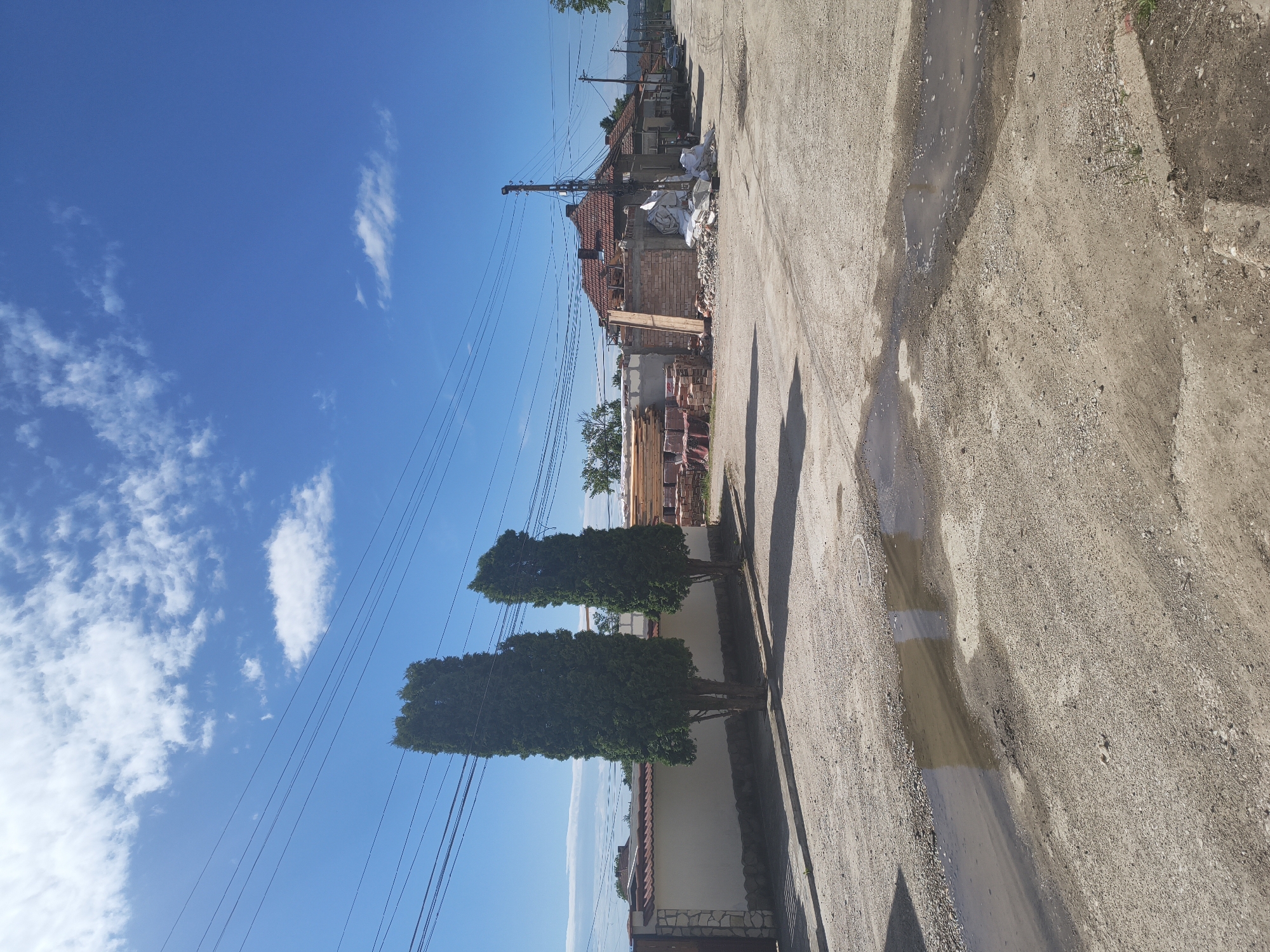 The image depicts an outdoor scene in Dolna Oryahovitsa, Bulgaria. A narrow, unpaved road dominates the foreground, showing signs of recent rain or standing water. The road is bordered by a building with a white wall and a red-tiled roof behind two tall, green, tree-like bushes. Beyond the building, construction materials are stacked up, next to an under construction house, implying ongoing construction work. Overhead power lines cross the blue sky with scattered white clouds.