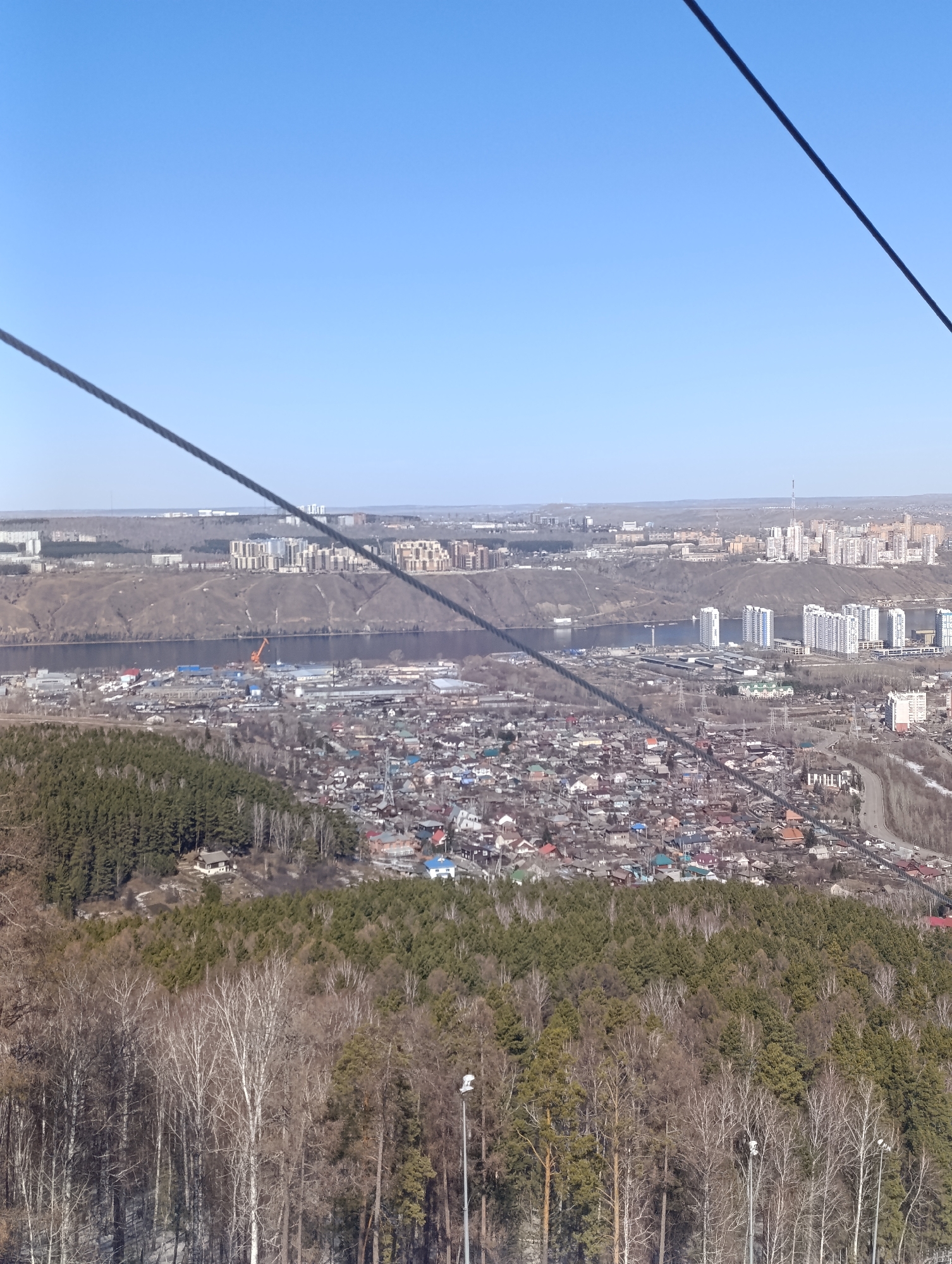 The image shows a panoramic view of Krasnoyarsk, Russia, from a high vantage point, likely a cable car or elevated structure. The sky is clear and blue. A large river runs through the scene, separating different parts of the city. On the left side of the river, there is a mix of industrial areas and low-rise buildings. On the right side, there are taller apartment buildings and residential areas. The foreground consists of dense forests with trees in varying stages of foliage. A cable car wire runs across the upper corners of the image.