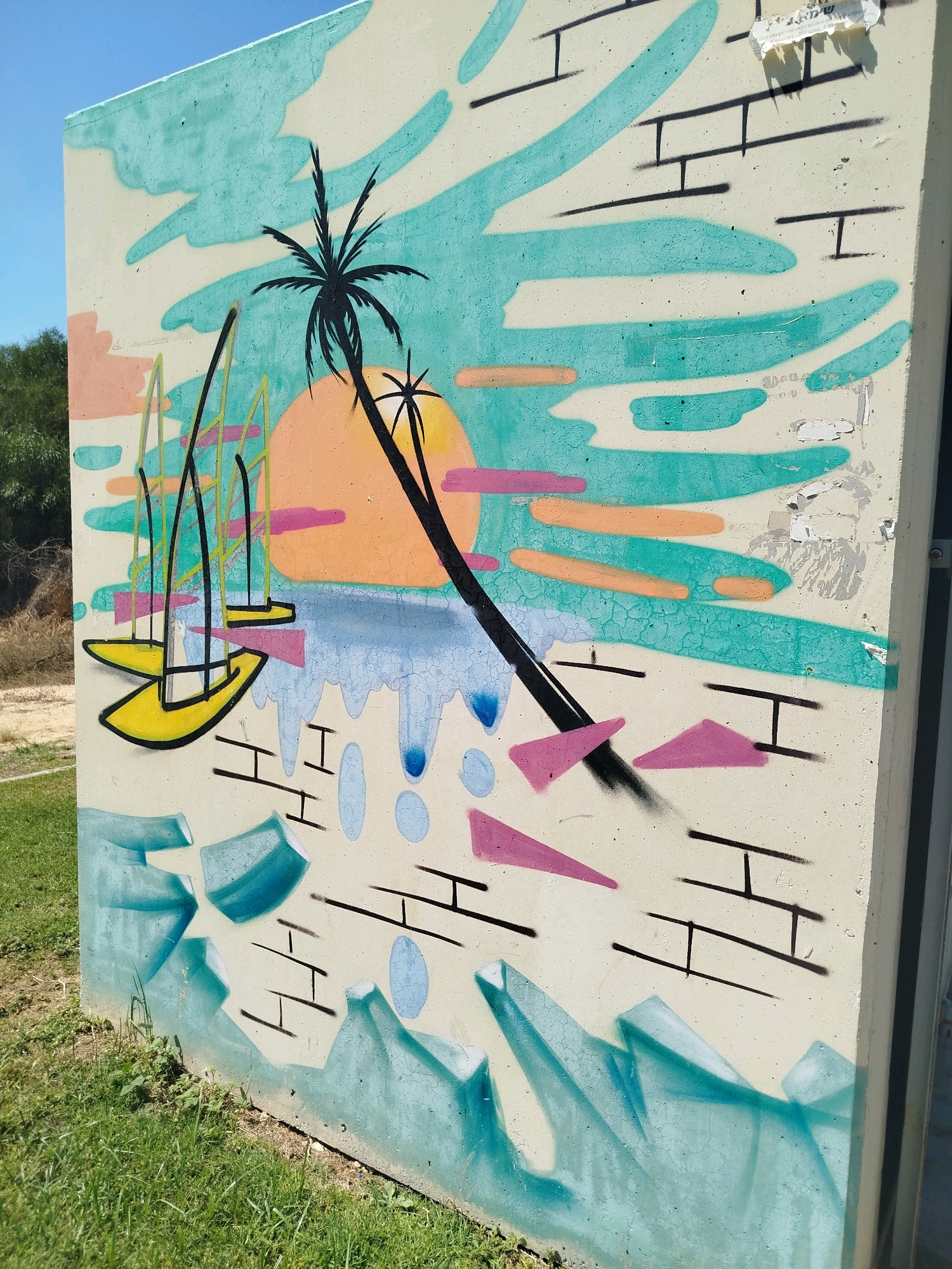 The image shows a concrete utility box mural painted with a vibrant, stylized beach scene. The mural features a large, orange sun setting over a blue ocean with dripping water effects. A silhouette of a leaning palm tree dominates the center. In the foreground, there are yellow sailboats with black masts, and abstract pink and teal shapes scattered throughout. The background is a pale yellow sky with teal cloud-like forms and horizontal orange lines. The mural is located outdoors, with green grass and some dry brush visible in the lower portion of the frame. The concrete surface of the utility box shows some signs of wear and tear, with peeling paint and cracks. The overall style is reminiscent of retro-futuristic or synthwave aesthetics. The location is identified as Ashkelon, Israel, suggesting this is a public art installation within the city.