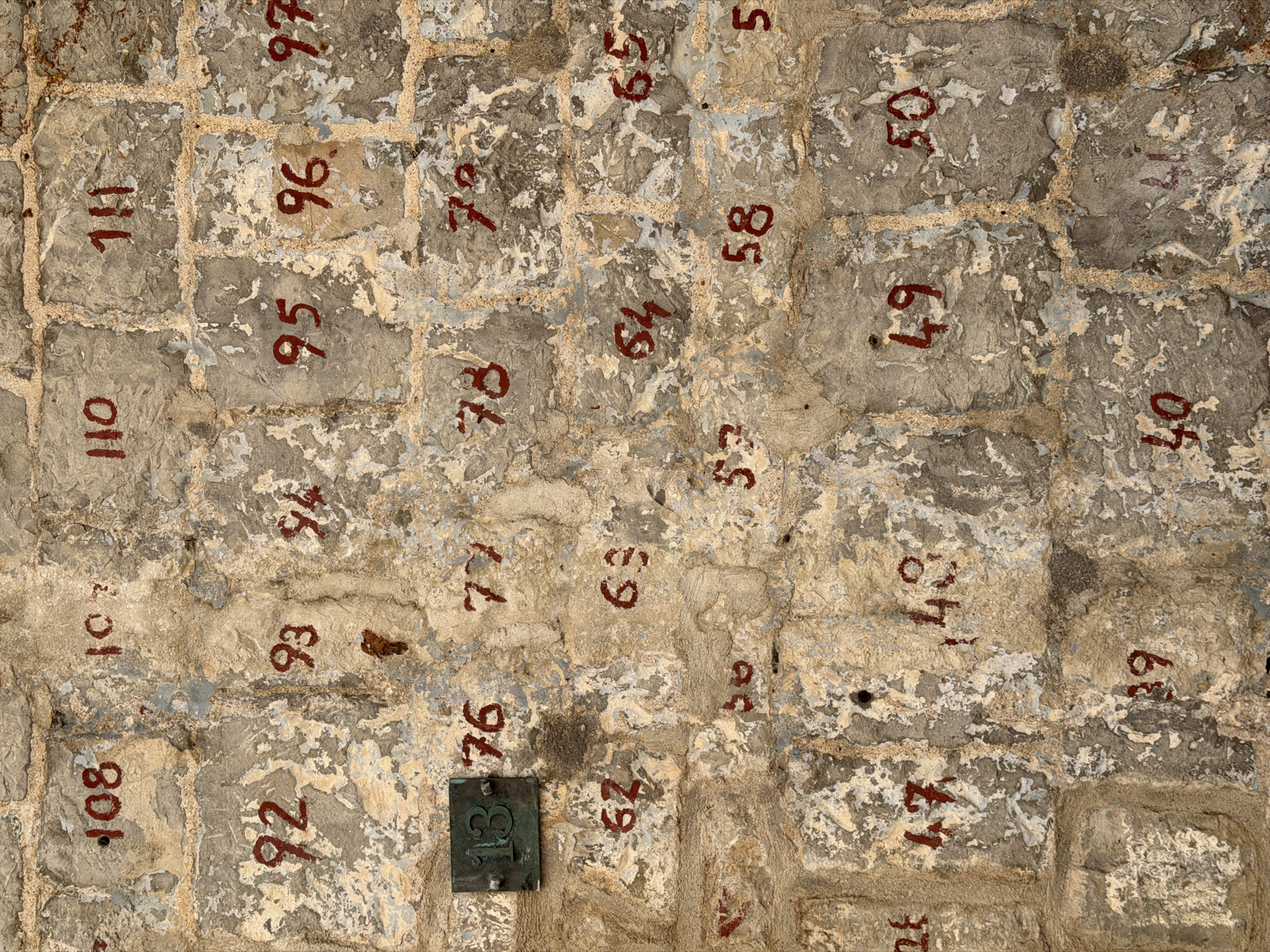 The image shows a section of a weathered stone wall in Budva, Montenegro, with red numerals painted on individual stones, presumably numbering apartments or houses. The numbers range from 108 down to 39 and 40, indicating a sequence. Some numbers are clear, while others are faded or partially obscured by the rough texture and discoloration of the stone. A small, square metal plaque with the number "13" in green is also visible, attached to one of the stones. The overall appearance suggests an old, possibly historical, part of the city.