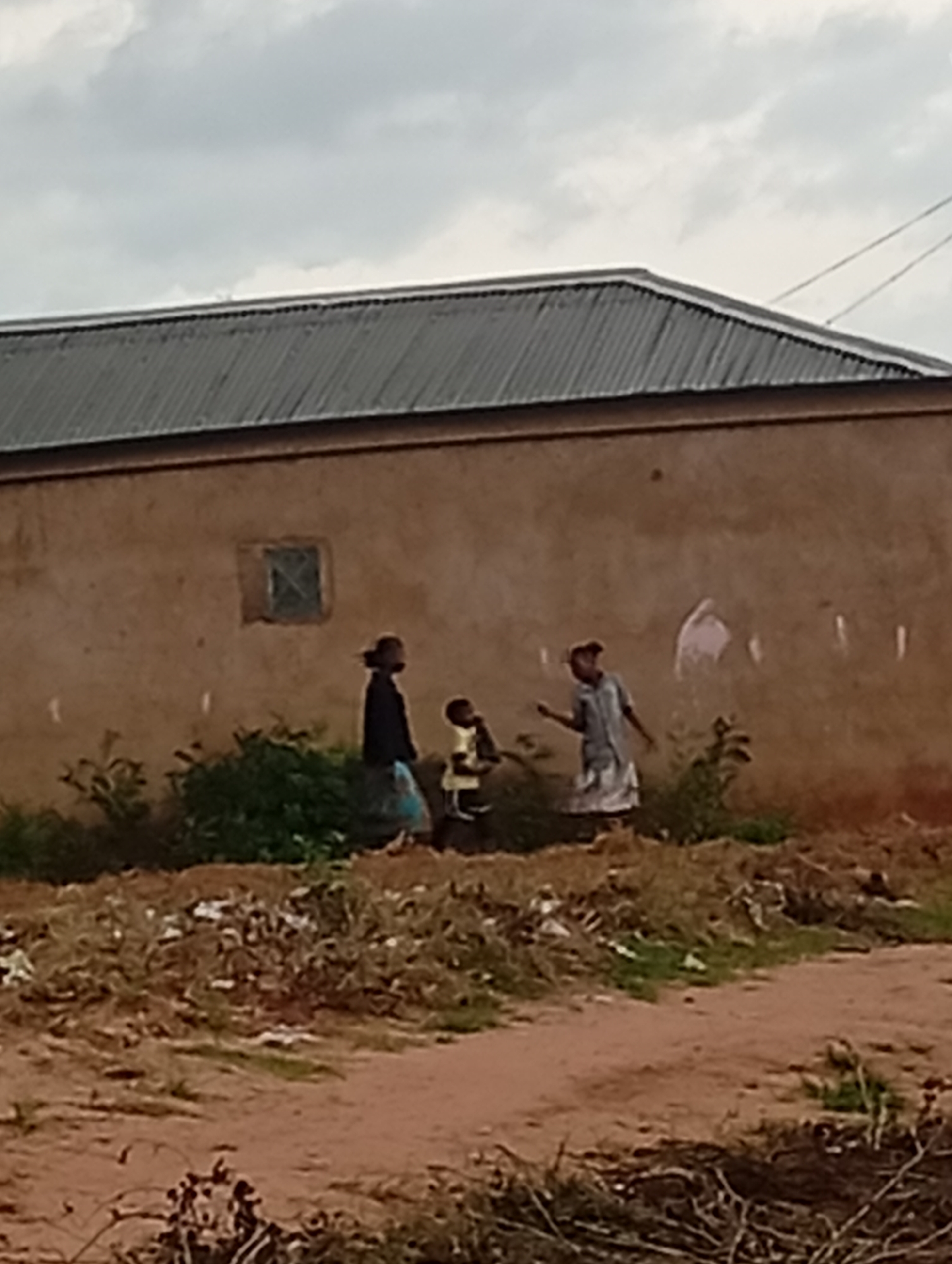 The image depicts an outdoor scene in Tumfure, Nigeria, with a focus on a simple, unplastered building with a corrugated metal roof. In front of the building, three individuals, appearing to be children, are visible. One child, wearing a dark top and a blue skirt, stands to the left. In the center, a smaller child in a yellow and black striped top is positioned. To the right, a child in a light-colored dress is engaged in what appears to be an interaction with the other two. The ground in front of the building is covered with debris and dry vegetation, suggesting an unkempt or undeveloped area. The sky is overcast with clouds. The overall impression is a candid shot capturing a moment of everyday life in a rural or semi-urban environment.