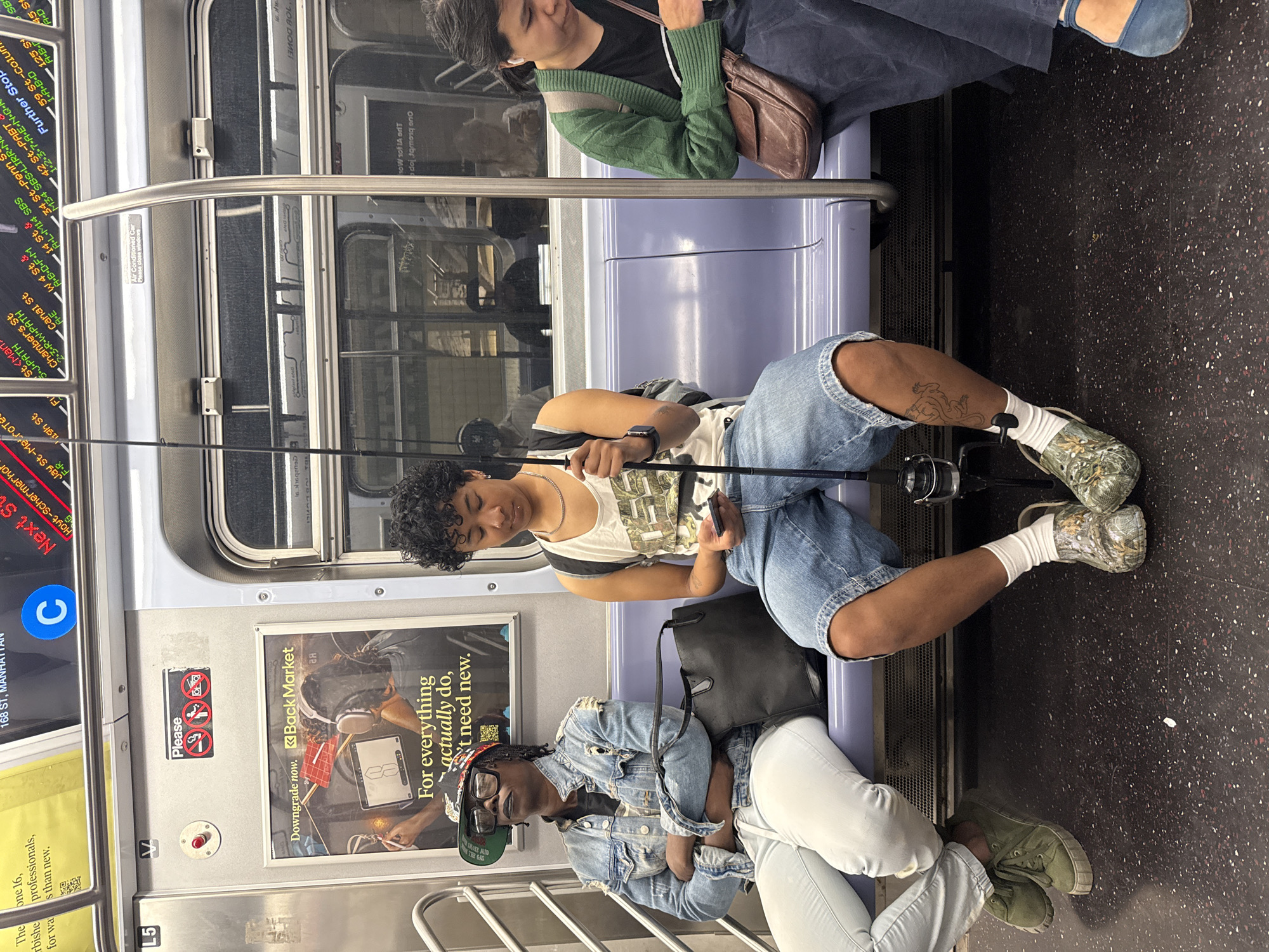 The image shows the interior of a New York City subway car. A passenger sits with a fishing rod resting across their lap. Another passenger sits at the edge of the seat. A poster on the wall advertises Back Market, a company that sells refurbished electronics. On the left side of the image, a digital screen displays subway routes, including the C line to 168 St, Manhattan. The current station listed is Hoyt-Schermerhorn St. Above this, several stations are shown on different lines. The floor is a dark color with speckled dots.