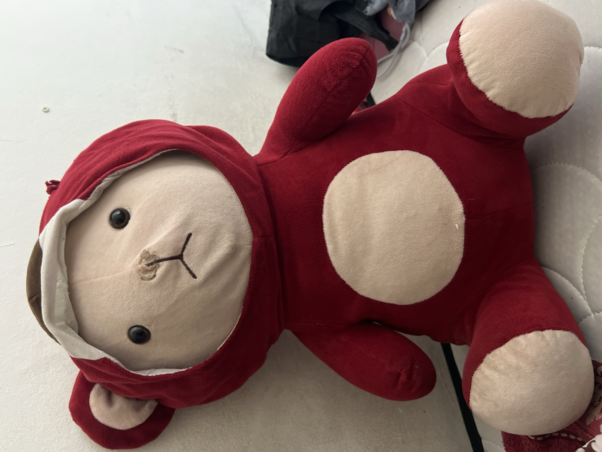 A plush toy bear, predominantly red with a lighter tan stomach and paws, is seated against a white wall. The bear wears a red hooded costume with a white lining. Its eyes are black and round, and its nose and mouth are stitched with brown thread. The bear's left ear is visible, with a tan inner ear. The background includes a portion of a dark fabric item to the right of the bear. The scene is indoors, likely a bedroom, and appears to be a casual setting. The location in Denpasar, Indonesia, is not evident from the visual information provided.