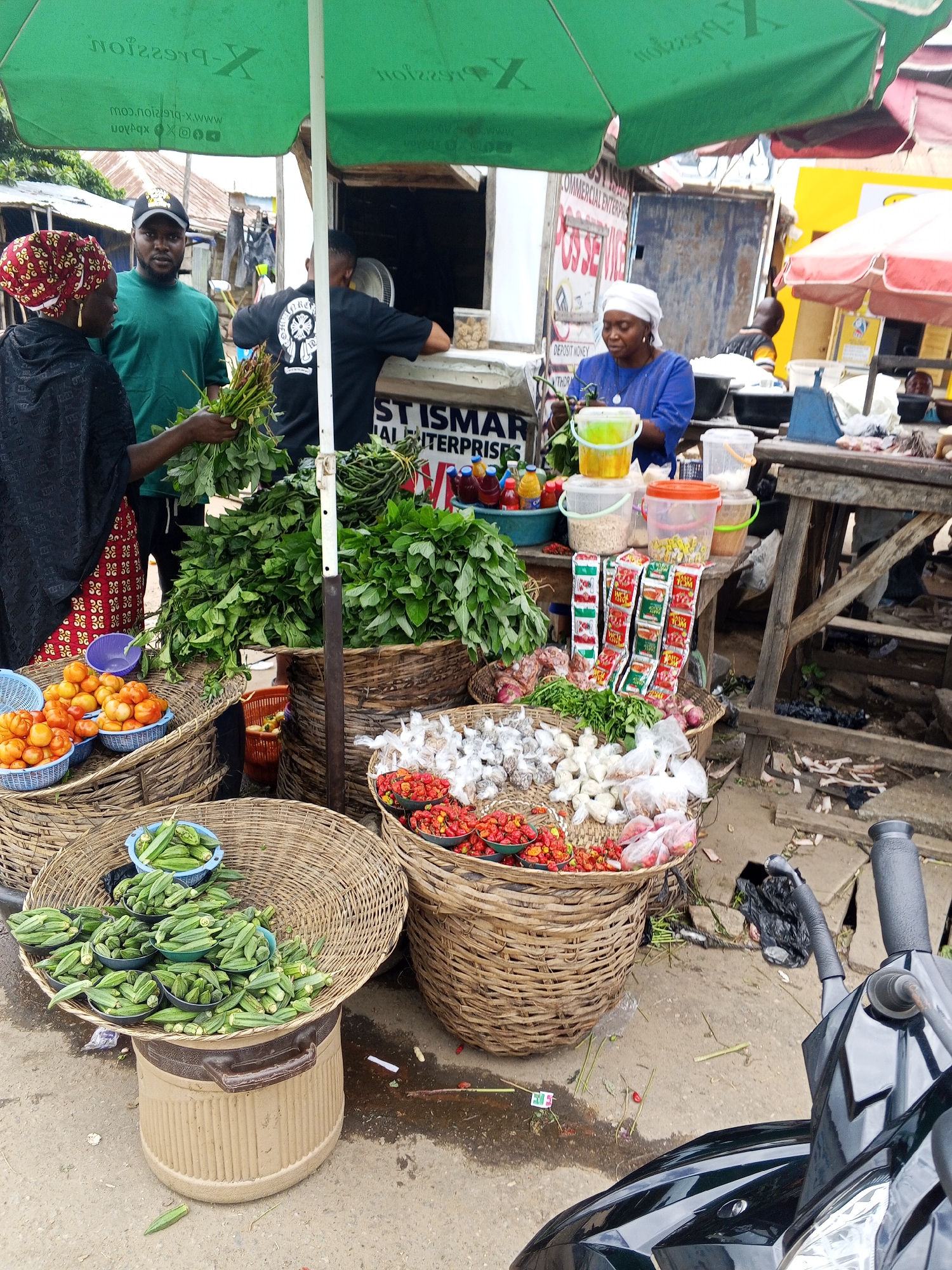The image depicts a vibrant street market scene in Zango-ndaji, Nigeria, under a large green canopy. In the foreground, a woman wearing a red and yellow headscarf and a black abaya holds a bundle of green leafy vegetables. To her left, a man in a green t-shirt and a baseball cap looks at the camera. Behind them, another person in a black t-shirt is visible.
The market stalls are laden with fresh produce. Large woven baskets overflow with tomatoes, okra, and chili peppers. Other baskets contain garlic and onions. Several plastic containers hold dry goods, and rows of small, colorful packets, likely spices or seasonings, are displayed prominently. Bottles of what appear to be beverages are also visible on a wooden table. The ground is a mix of sand and wet patches, indicative of recent rain or water usage in the market. In the lower right corner, a portion of a black motorcycle is visible, suggesting a mode of transportation within or to the market. The overall atmosphere is one of bustling commerce and the everyday life of a Nigerian market.