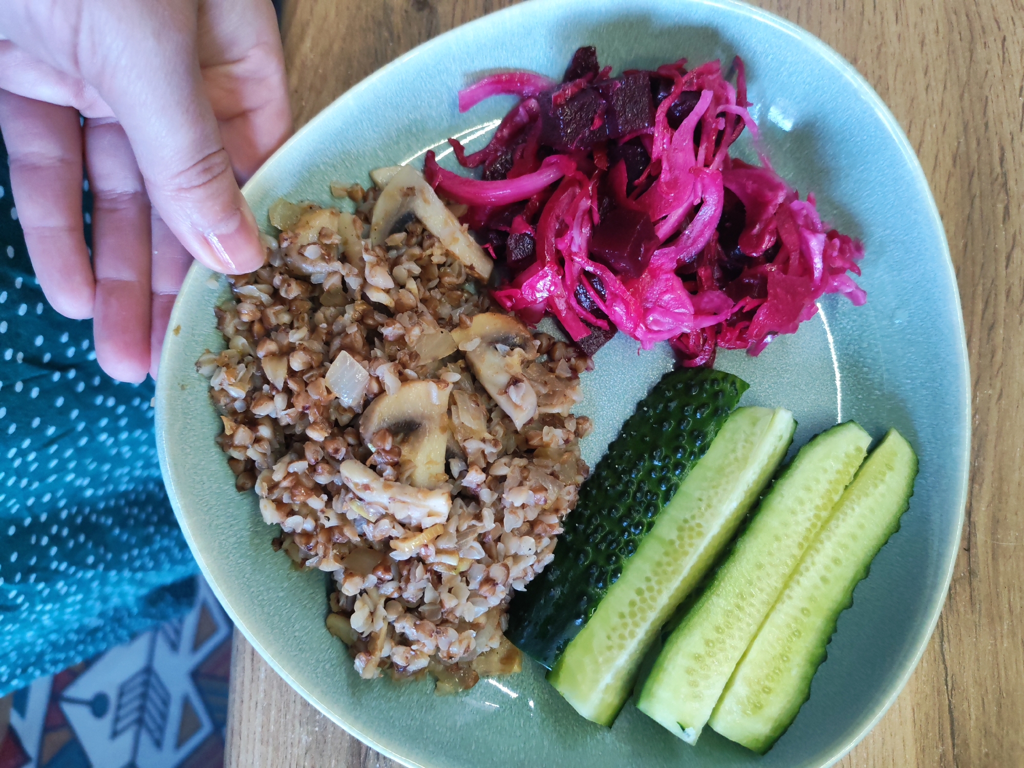 An overhead, close-up shot displays a meal served on a light blue-green ceramic plate, held by a person's hand on the left. The person is wearing a dark green garment with white polka dots.
The plate holds three distinct food items. On the left side, there is a portion of cooked buckwheat (kasha) mixed with chopped onions and sliced mushrooms. In the upper right section, a vibrant pile of intensely pink and purple pickled vegetables, likely consisting of shredded cabbage and cubed beets, is visible. The lower right portion of the plate features three pieces of cucumber: one whole gherkin-like cucumber and two longitudinally sliced halves.
The meal is presented on a light brown wooden table. A segment of a patterned rug or mat with geometric designs in shades of blue, orange, and white is partially visible in the bottom left corner, beneath the person's arm. The scene depicts a casual indoor setting, possibly a home, consistent with the context of Izhevsk, Russia. No text is visible in the image.
