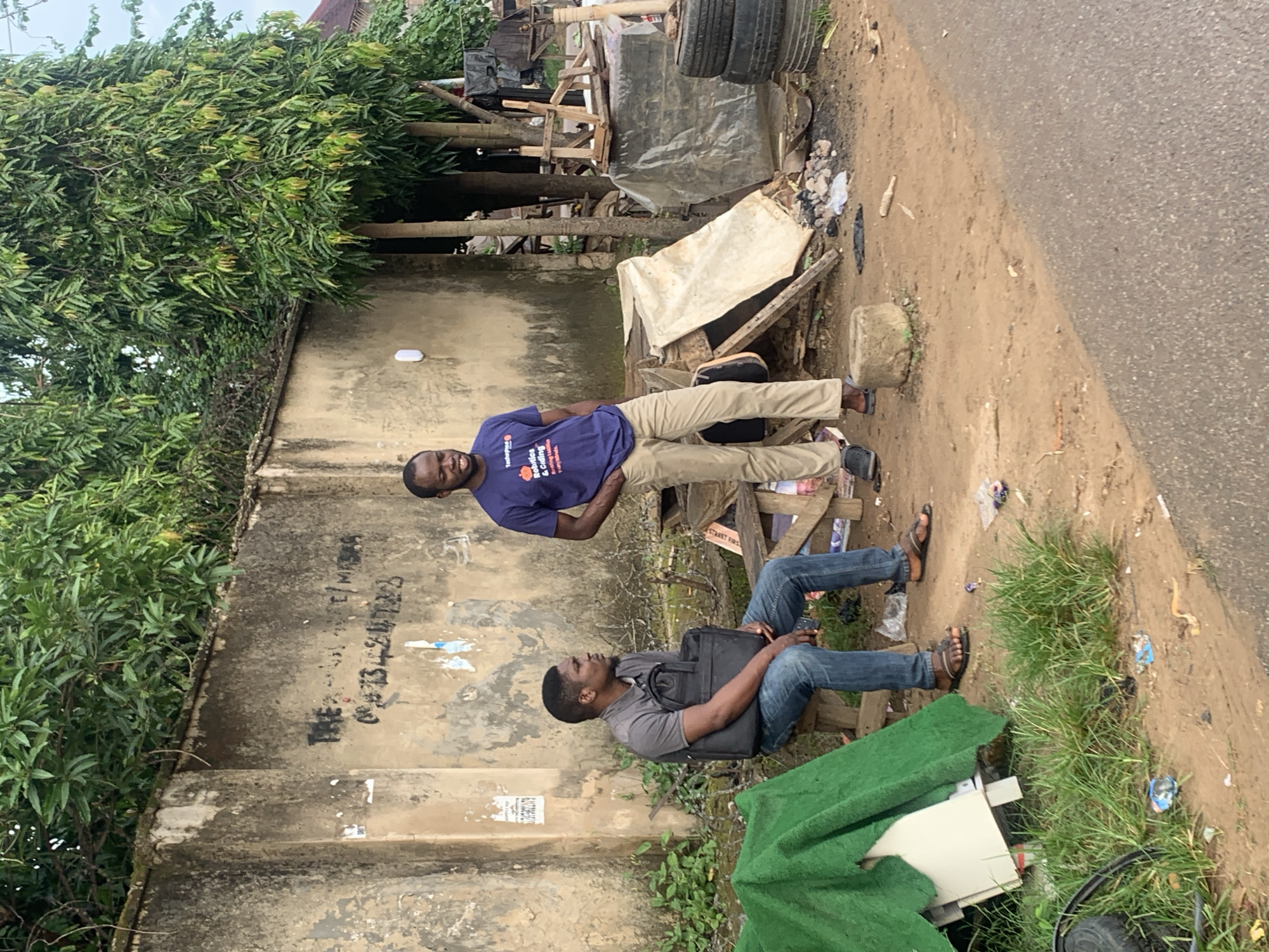 Two men are standing and sitting by a concrete wall with graffiti in an urban environment, possibly Abuja, Nigeria. The man sitting on a wooden bench is wearing a grey polo shirt and blue jeans, holding a black laptop bag. The man standing is wearing a blue t-shirt with an orange graphic and khaki pants. Behind them, there is a wall with faded writing, overgrown green vegetation, and debris including tires, wood, and white tarps. A green tarp covers some objects to the left of the bench. The ground is dusty and uneven, with some scattered trash.