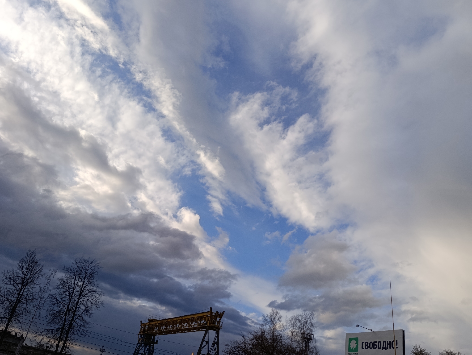The image captures a dramatic sky over Chaikovsky, Russia, dominated by an expansive cloud formation. White, grey, and dark grey clouds create dynamic patterns across the frame, interspersed with distinct patches of clear blue sky, particularly in the upper center. The varied lighting on the clouds suggests either an approaching or receding weather system, or the sun breaking through an otherwise overcast sky.
In the lower left, silhouettes of several bare deciduous trees are visible against the dark clouds, indicative of winter or early spring. Below them, what appear to be power lines stretch horizontally. A prominent yellow-painted industrial structure, likely a large gantry crane or similar heavy machinery, occupies the lower center, showcasing its intricate truss framework.
On the lower right, a white billboard is partially visible. It features a green, four-leaf clover-like logo on the left side. The main text on the billboard reads "СВОБОДНО!", which translates from Russian to "FREE!" or "AVAILABLE!". Below the logo, smaller text says "грин медиа" (Green Media).
The overall lighting suggests either a late afternoon or early evening setting, or a day with heavy cloud cover allowing some sunlight to penetrate. No human figures or direct activity are visible; the scene is static, focusing on the skyscape and the urban/industrial elements beneath it.