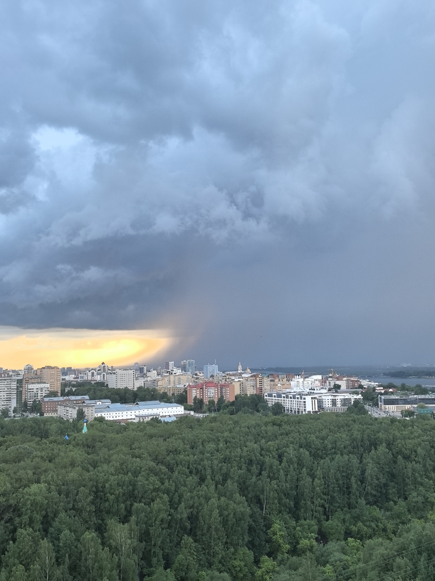 This is an aerial view of a city skyline in Perm, Russia, under a dramatic sky with storm clouds. In the foreground, a dense forest of green trees occupies the lower half of the image. Behind the forest, a city with a mix of mid-rise and high-rise buildings is visible. The buildings are predominantly modern, with many white facades and varied architectural styles.
The sky is characterized by dark, heavy storm clouds dominating the upper portion of the frame. To the left, a band of bright, yellowish light breaks through the clouds, possibly indicating sunlight or a clearing storm. A faint rainbow or light shaft can be observed within this brighter area. The contrast between the dark clouds and the bright light creates a striking visual effect.
The image captures a sense of an impending or passing storm over the urban landscape and the natural environment of the forest. No people are discernibly present in the image. The overall impression is one of a powerful natural phenomenon interacting with the built environment.