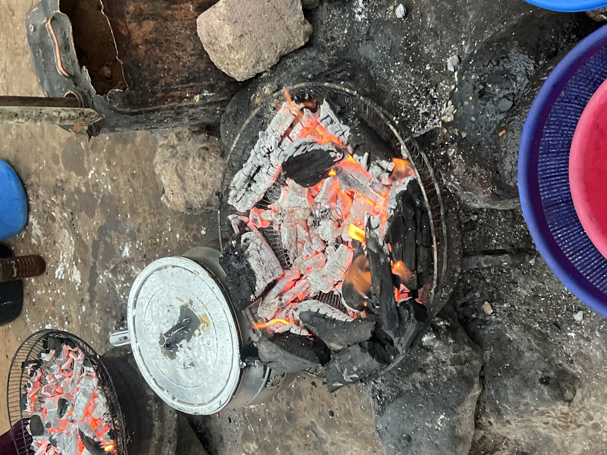 This image captures a rustic outdoor cooking setup, likely in Gura Top, Nigeria. The scene is dominated by two charcoal grills, suggesting meal preparation.
In the foreground and center-right, a larger, round charcoal grill contains actively burning charcoal, exhibiting bright orange and red embers amidst pieces of black charcoal and white ash. This grill appears to be a metal grate set over a base. To its left, a smaller charcoal grill also holds glowing embers, though these appear more covered in white ash.
Positioned between these two grills is a round, metallic pot with a silver lid, resting on what appears to be a smaller, simple stove or directly on the embers of the left grill. Several rough-hewn stones are scattered around the cooking area, potentially serving as supports or part of the cooking structure.
In the upper left corner, a dark, rusted metal object, possibly a cut-open container or part of a makeshift stove, is visible. To the right of the main grill, parts of two colorful plastic bowls, one blue and one red, are seen, hinting at dishes or ingredients.
The ground surface is uneven, consisting of dirt or concrete, and is heavily speckled with white ash and dark charcoal dust, indicating an area frequently used for cooking with open fires. The overall setting is simple and functional, typical of an outdoor or semi-sheltered cooking space. There are no people visible, and no discernible text. The warm glow of the coals suggests an active cooking session, but the time of day or specific weather conditions are not discernible from the image.