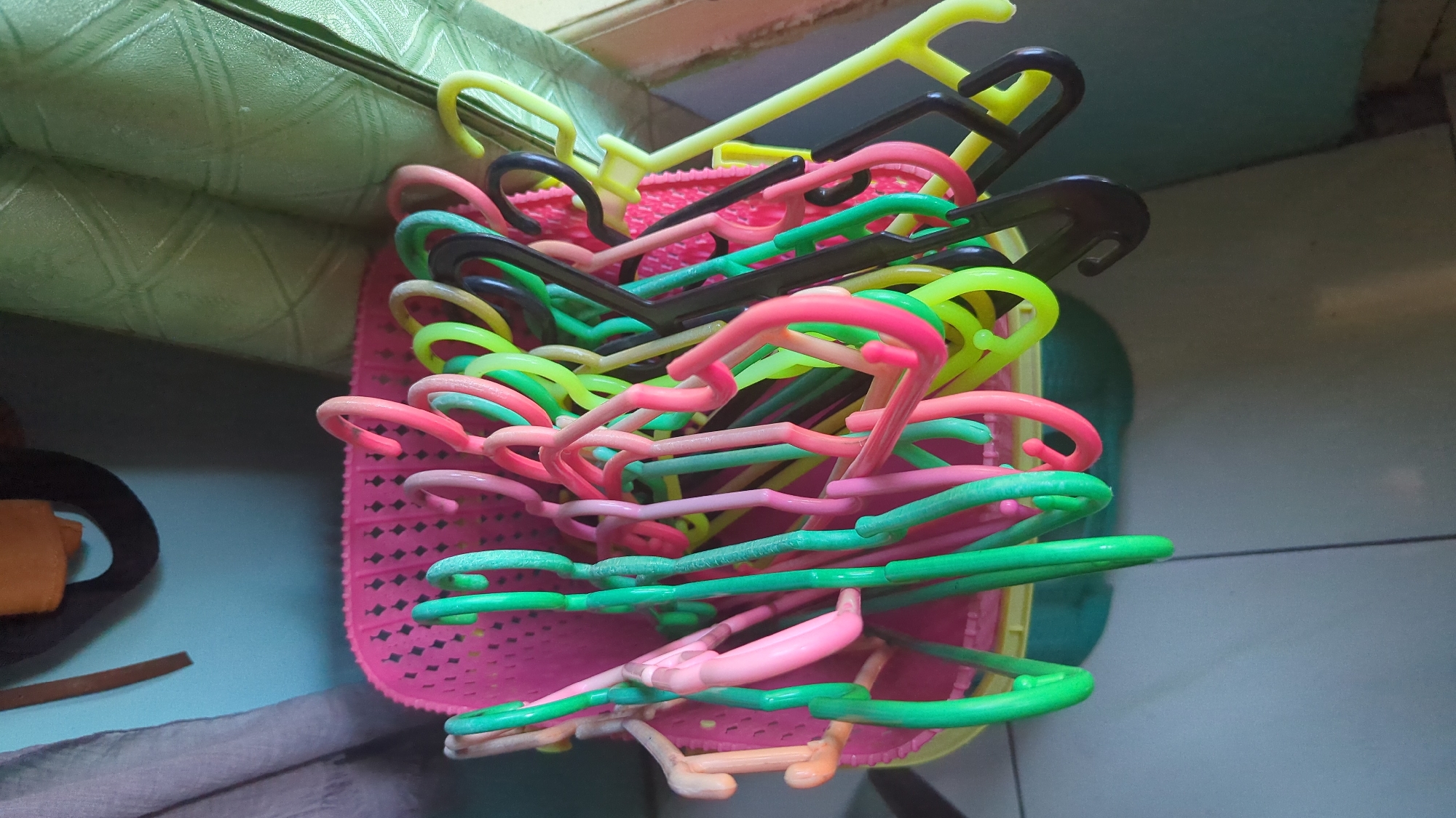 A close-up, high-angle shot shows a collection of colorful plastic clothes hangers overflowing from a pink woven plastic basket. The hangers are a mix of pink, green, yellow, and black. Some hangers are stacked inside the basket, while others spill out over the sides, with their hooked ends dangling. The basket is sitting on a tiled floor. In the background, a light green textured curtain hangs to the right, and a light blue wall is visible to the left, with a dark object hanging on it. The scene appears to be indoors, possibly in a domestic setting.