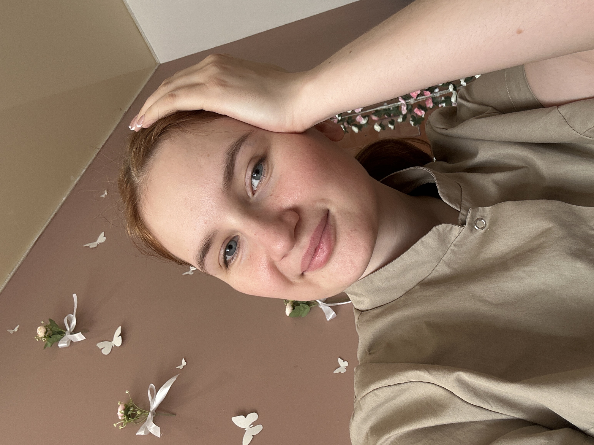 A close-up photograph shows a young woman with light skin, blue eyes, and reddish-brown hair. She is smiling slightly and her hand is resting on her forehead. She is wearing a light brown shirt. The background is a brown wall decorated with small white paper butterflies and floral arrangements tied with white ribbons. The photograph might be taken in Chaikovsky, Russia, though this is not possible to confirm from the image alone.