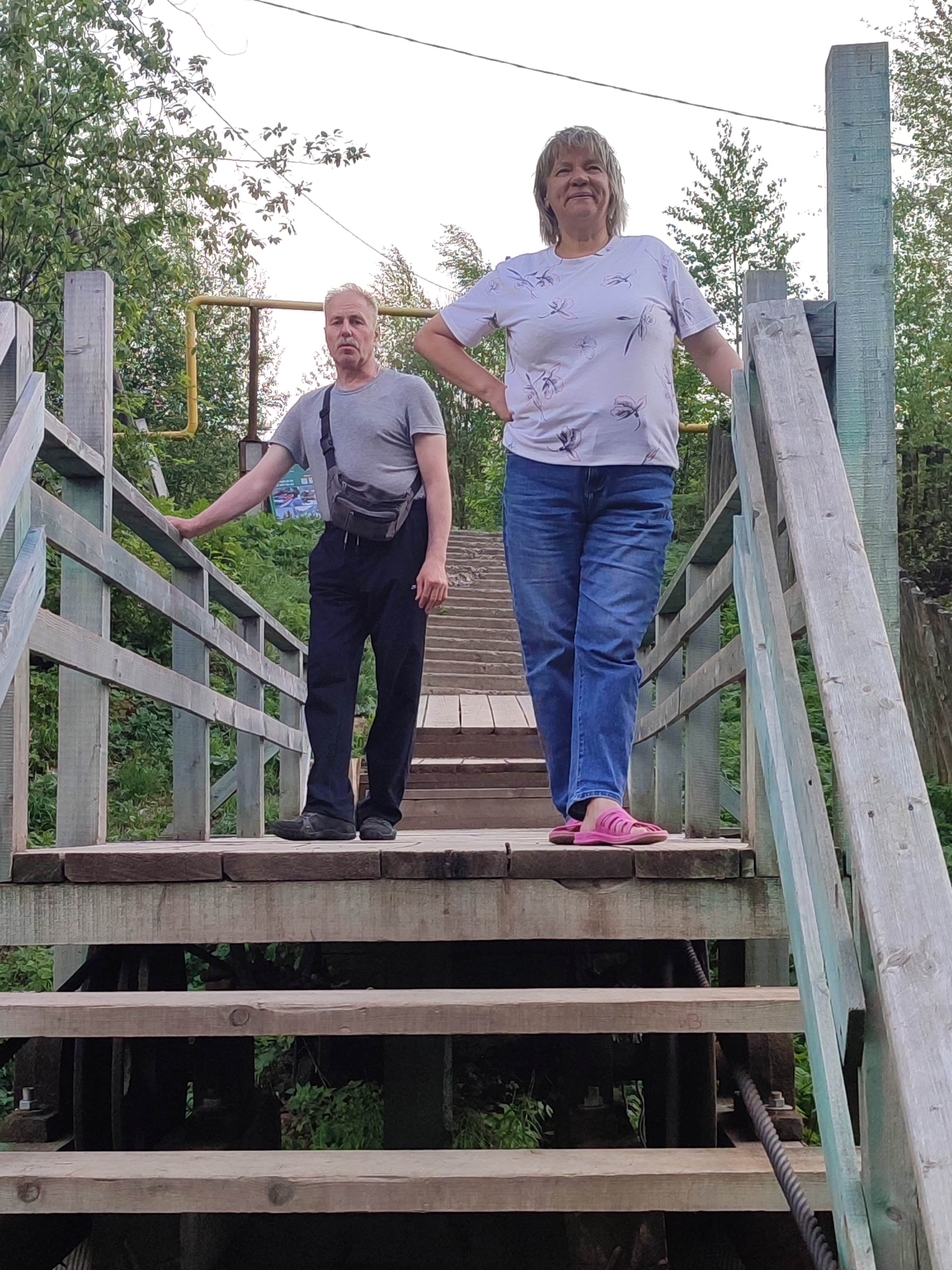 Two adults stand on a wooden stairway structure in Усьва, Russia. A man with short blond hair and a gray t-shirt stands on the left, leaning on the wooden railing with his left arm. He wears dark pants and a small gray bag across his torso. A woman with short blond hair and a white t-shirt with a floral pattern stands to the right of the man. She wears blue jeans and pink sandals. Both people look forward. The stairs lead downwards. Dense vegetation surrounds the structure, and a yellow gas pipe is visible on the left.