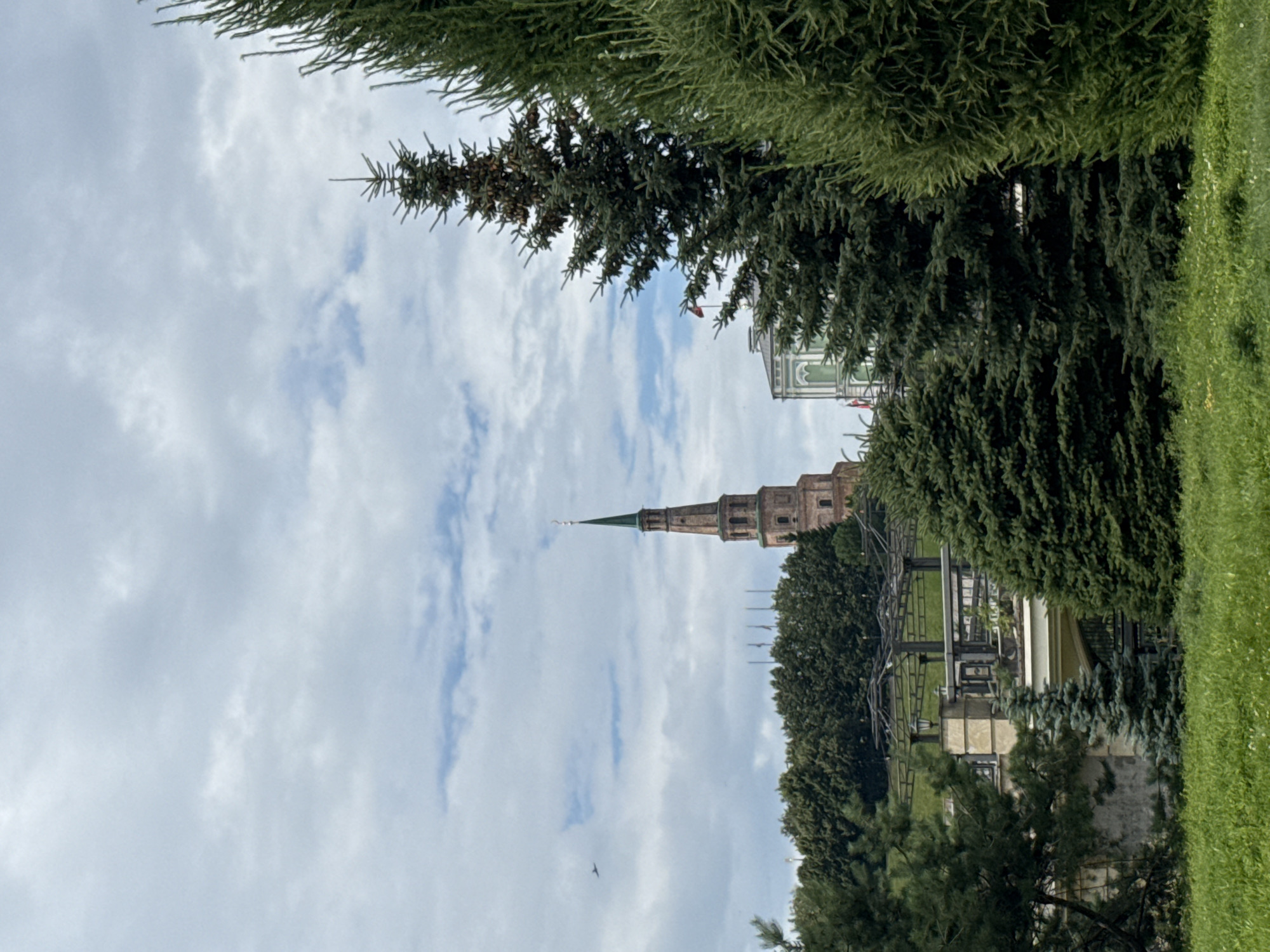 The image captures a view of the Suyumbike Tower in Kazan, Russia, from a park-like setting. The tower, a prominent red-brick structure with a green spire, stands tall against a cloudy sky. In the foreground, lush green grass and various coniferous trees, including several tall fir trees, dominate the scene. A portion of a building with a green roof and white architectural details is visible to the right of the tower. In the far left of the sky, a small airplane is visible. The overall impression is of a serene, natural environment offering a glimpse of historical architecture.