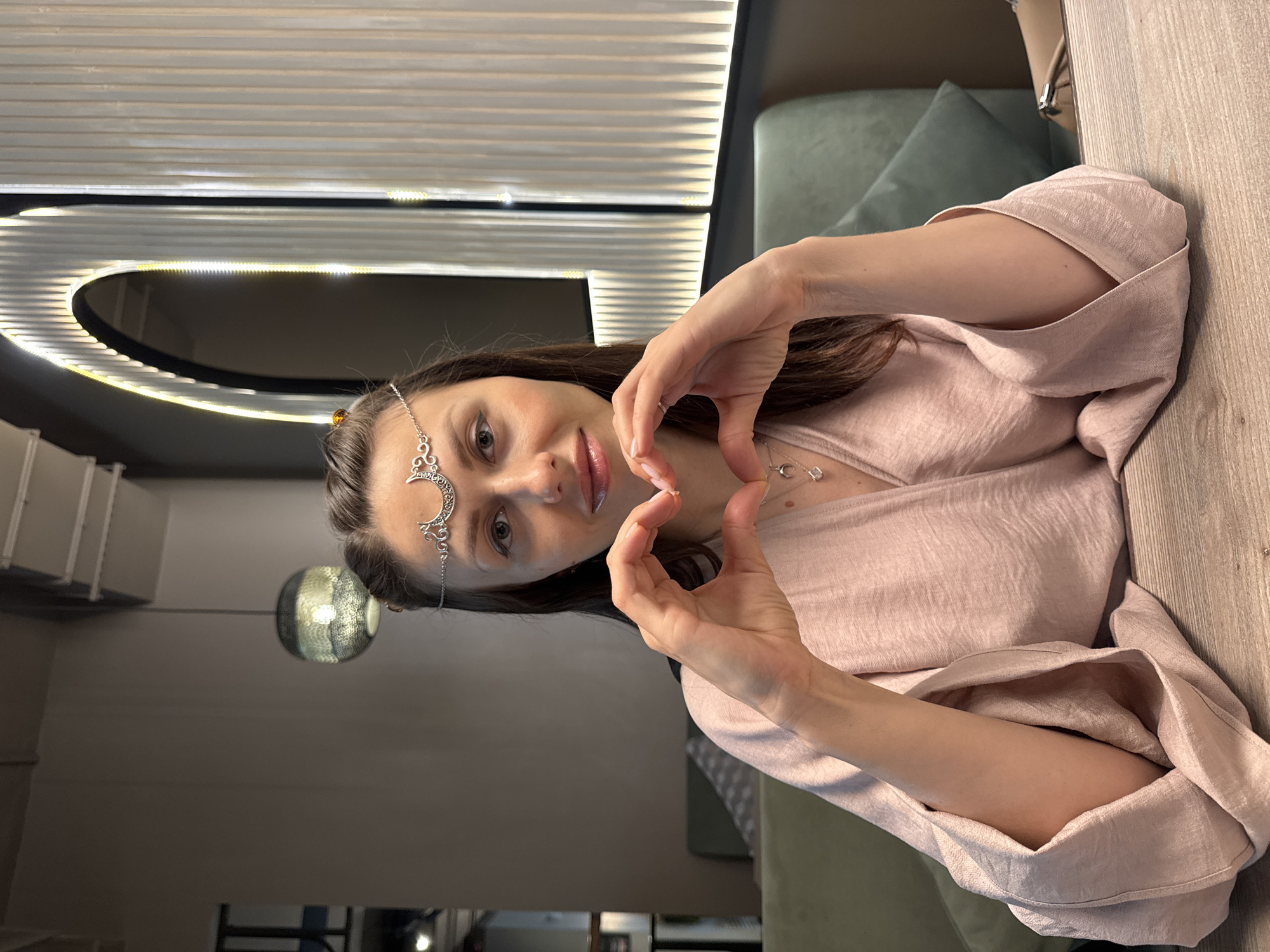 A woman is indoors, presumably in Unknown, Russia. She is fair-skinned with brown hair and is wearing a decorative silver headpiece, a light pink blouse, and a necklace with small, square pendants. She is smiling and making a heart shape with her hands. Behind her, there is a wall with horizontal lines and an oval-shaped light fixture. A green pillow is visible in the background, as is a light-colored wooden table. A spherical lamp is partially visible in the background on the left side of the image.