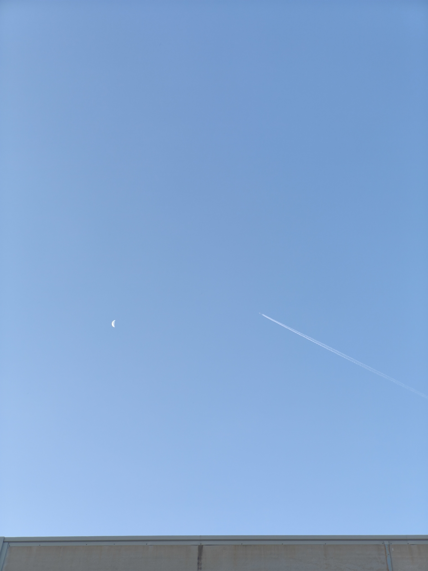 A clear blue sky dominates the upper portion of the image, with a crescent moon visible on the left side and a contrail from an airplane streaking across the right side. The lower portion of the image features a concrete barrier or wall, suggesting an outdoor or industrial setting. The image appears to be taken in Nàquera, Spain. There are no people or animals visible in the scene.