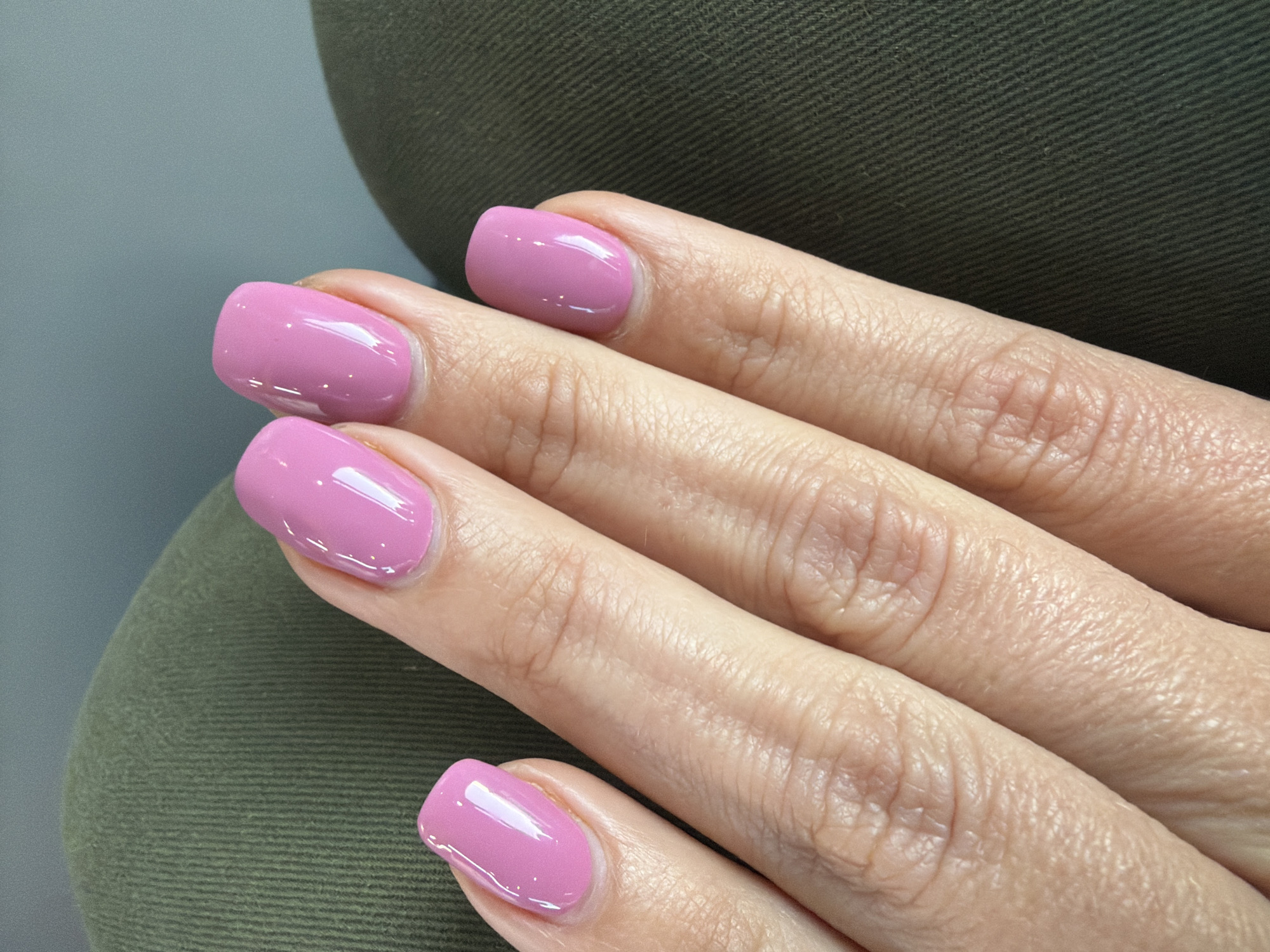 A close-up studio shot shows the fingers of a human hand with a fresh, glossy pink manicure. The nails are short and square-shaped, reflecting light. The fingers are slightly wrinkled, suggesting age. The hand rests against a coarse, green fabric background. No location is identifiable from the image.