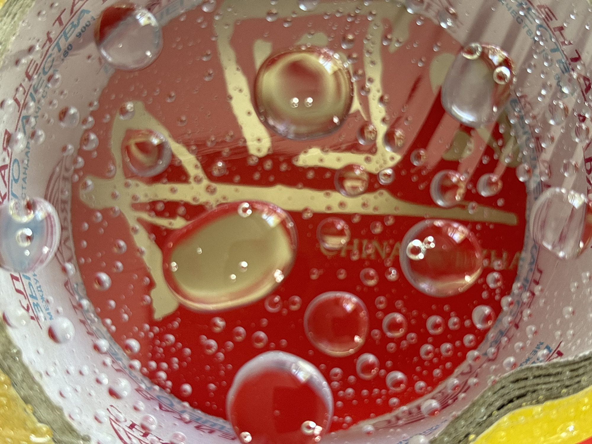 The image shows a close-up, top-down view into a container with a red and white interior. Numerous water droplets of various sizes cover the surface. The red interior is decorated with gold-colored designs and the word "CHINA" is visible in one area. The container's edge is visible and appears to be made of cardboard. Text in Russian is visible around the interior edge of the container. The subject and environment strongly suggest a disposable food container, and the writing suggests its origin. No specific location in Chaikovsky, Russia can be determined from the image.
