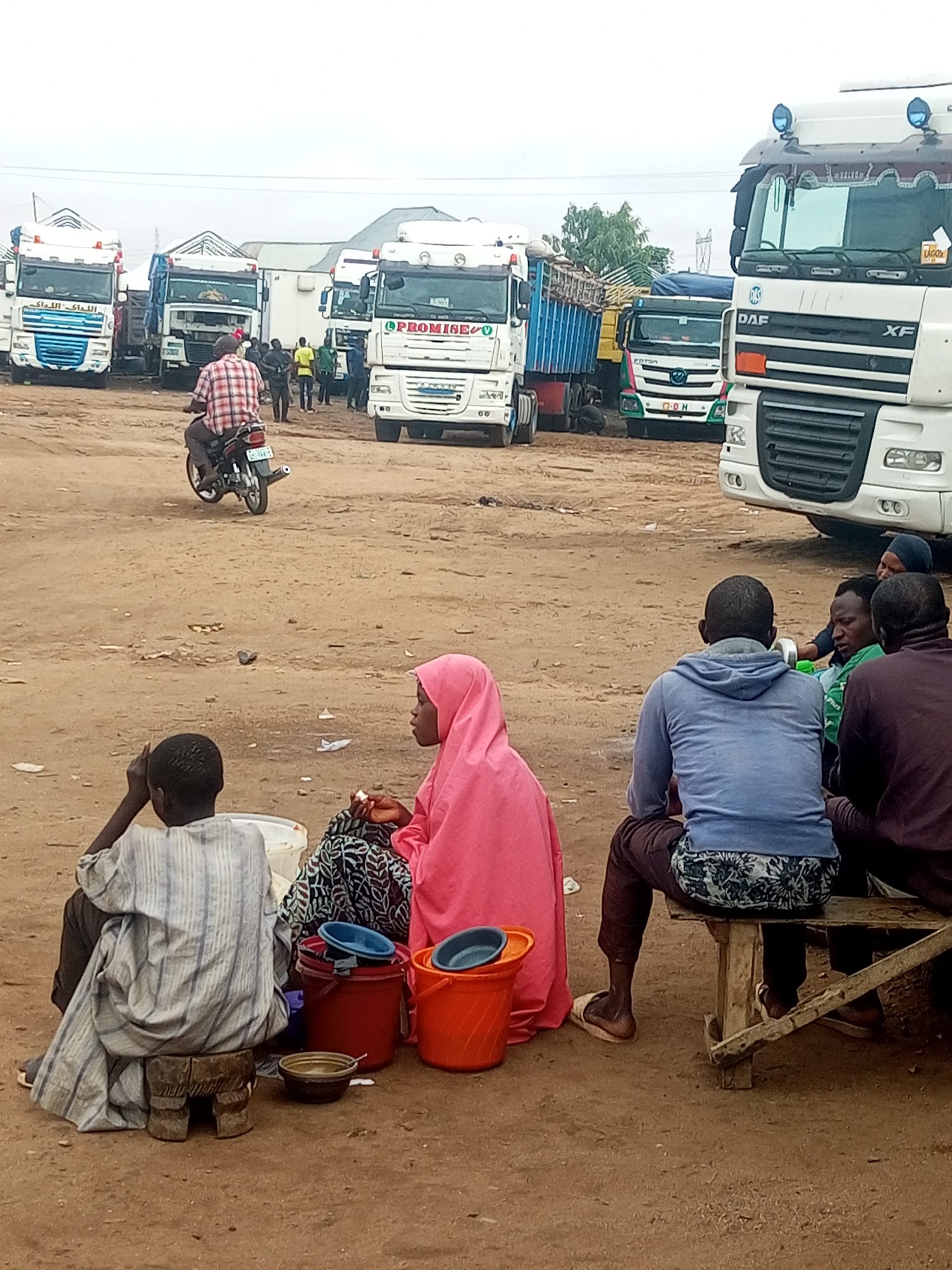 This image captures an outdoor scene at a truck stop or unpaved commercial area in Unknown, Unknown, under an overcast sky. The ground is dry, dusty, and light brown, scattered with minimal debris.
In the immediate foreground, a group of individuals with dark skin are seated. On the far left, a person wearing a light-colored, vertically striped garment and dark pants sits on a small, carved wooden stool, facing right with their back to the camera. Next to them is a white plastic bucket. To their right, another individual, likely female, wears a pink hijab and a patterned skirt, seated low to the ground, facing left. Near them are several plastic buckets in red, orange, and blue, along with a small brown bowl containing liquid.
Further to the right in the foreground, four individuals are seated on a rustic wooden bench. The person closest to the camera wears a blue hoodie and patterned shorts over dark pants, with light-colored sandals. To their right, another person wears a dark maroon long-sleeved top and dark pants. Two more individuals are partially visible within this group; one is wearing a green shirt. All are seated with their backs to the camera.
In the mid-ground, a person wearing a red and white plaid shirt and dark pants rides a black motorcycle with a silver exhaust pipe from left to right across the dusty terrain.
The background is dominated by numerous large commercial trucks. From left to right:
1. A white semi-truck with blue accents and Arabic script on the grille ("اللداك اللاك") and its trailer.
2. A white semi-truck with a blue open-top trailer, marked with "PROMISE" on the cab.
3. A white refrigerated truck.
4. A green and pink/red Foton truck with "FOTON" and "O-D N" on its grille, carrying a blue tarp-covered load.
5. Partially visible on the far right, a prominent white DAF XF semi-truck with "DAF" and "XF" clearly visible on its grille.
Numerous other individuals are seen standing and moving amongst the parked and moving trucks, suggesting activity related to transportation or commerce. Sparse green foliage from a tree is visible in the mid-background. Distant structures with light-colored roofs are faintly visible on the horizon.