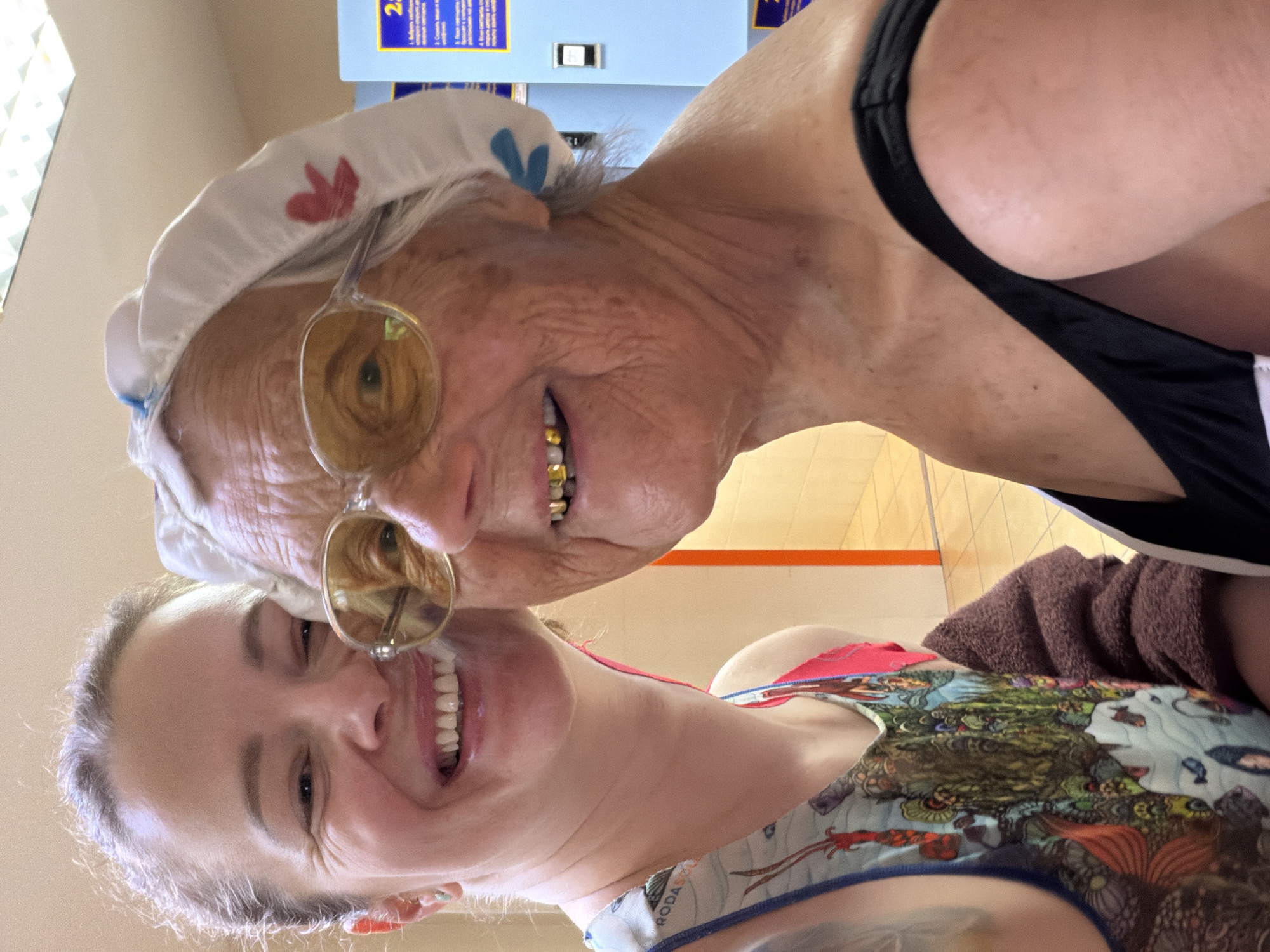Two people are close together, smiling at the camera. An older woman with a white swim cap adorned with red and blue floral patterns and gold-tinted glasses is at the top, and a younger woman wearing a patterned top is below. The older woman is wearing a black swimsuit and has gold teeth. The younger woman has light skin and is smiling broadly. In the background, there is a beige wall and floor, with a machine featuring blue and yellow signage. The location appears to be an indoor public space, possibly a pool or recreation center. There is a brown towel visible in the background as well. The city of Ждановіцкі сельскі Савет, Belarus is not apparent in the image.
