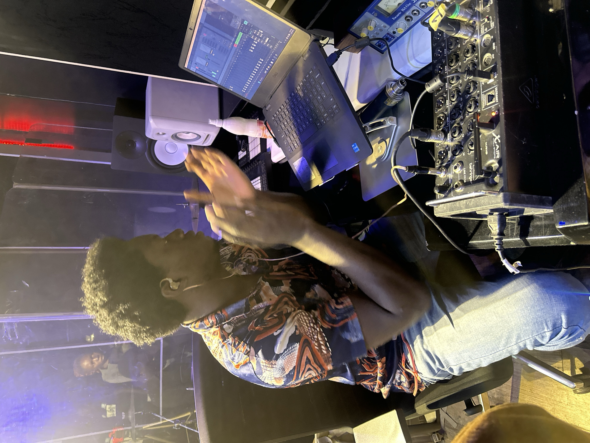 A Black male with dark, curly hair, wearing a colorful, patterned button-down shirt and light blue jeans, is seated at a desk in what appears to be a recording studio. He is wearing white earbuds and is looking towards a laptop that is open in front of him. His hands are raised and clapping, suggesting he is either demonstrating something or reacting to audio. The laptop screen displays a digital audio workstation interface.
Several pieces of audio equipment are visible on the desk: a black Behringer X AIR XR16 digital mixer, a blue and silver Focusrite ISA One analog preamplifier, a microphone, and various cables connecting the equipment. Two studio monitors, one black and one white, are positioned on a shelf behind the desk.
In the background, through a clear partition, a person with dark hair is visible, appearing to be in another area of the studio. Red lighting is visible in the upper background. The overall environment is dimly lit, with a focus on the audio equipment and the seated individual. The location is Barakin Lerawi, Nigeria.