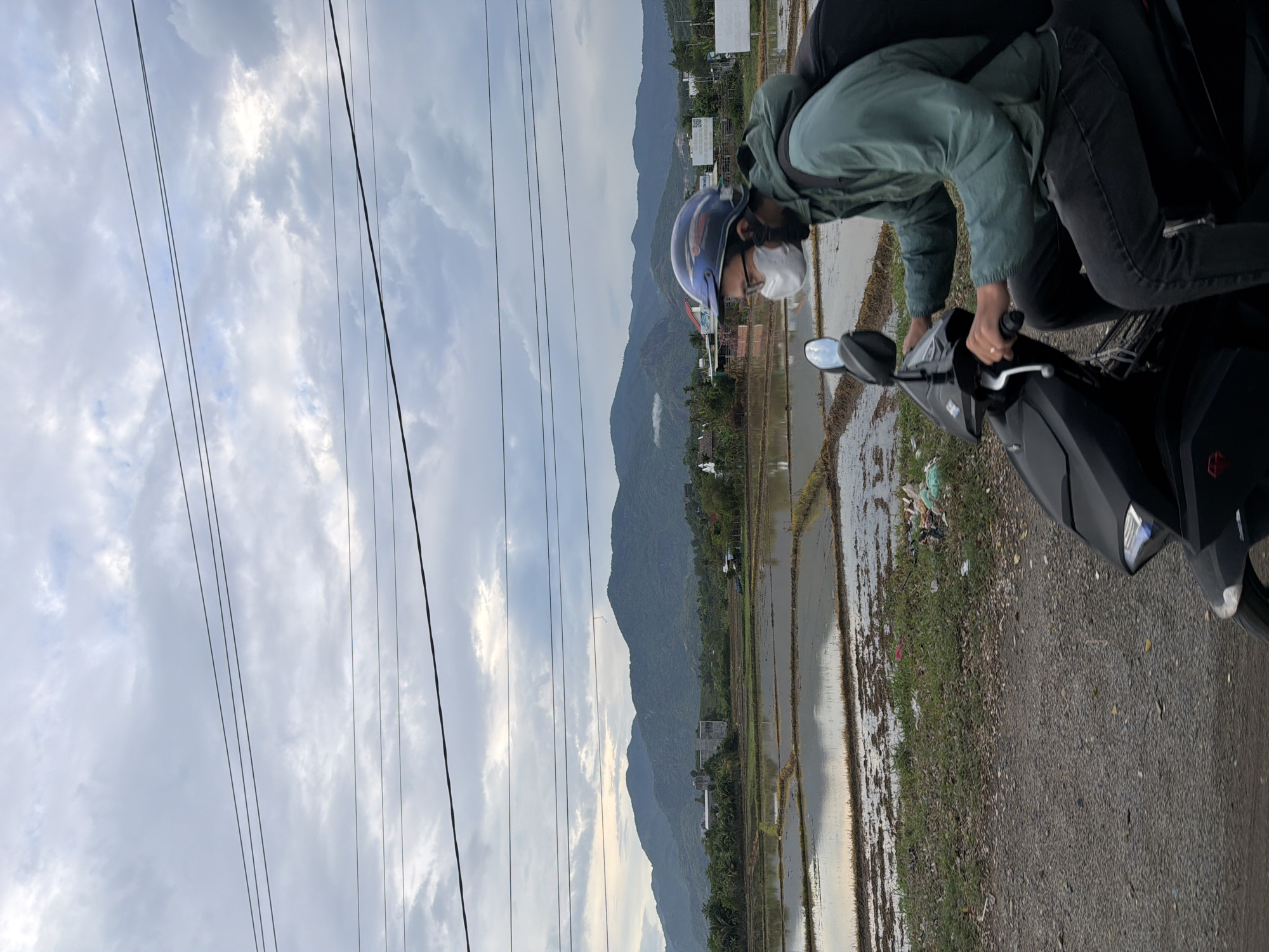 A person wearing a green jacket, dark pants, a helmet, and a face mask rides a black scooter through an agricultural area. The scooter has a side mirror and a reflector on the front. The person's right hand is on the handlebars. In the background, there are flooded rice paddies reflecting the cloudy sky. Beyond the paddies are green hills, a few buildings, and trees. Power lines are visible in the sky. The location is Diên Thọ, Vietnam.