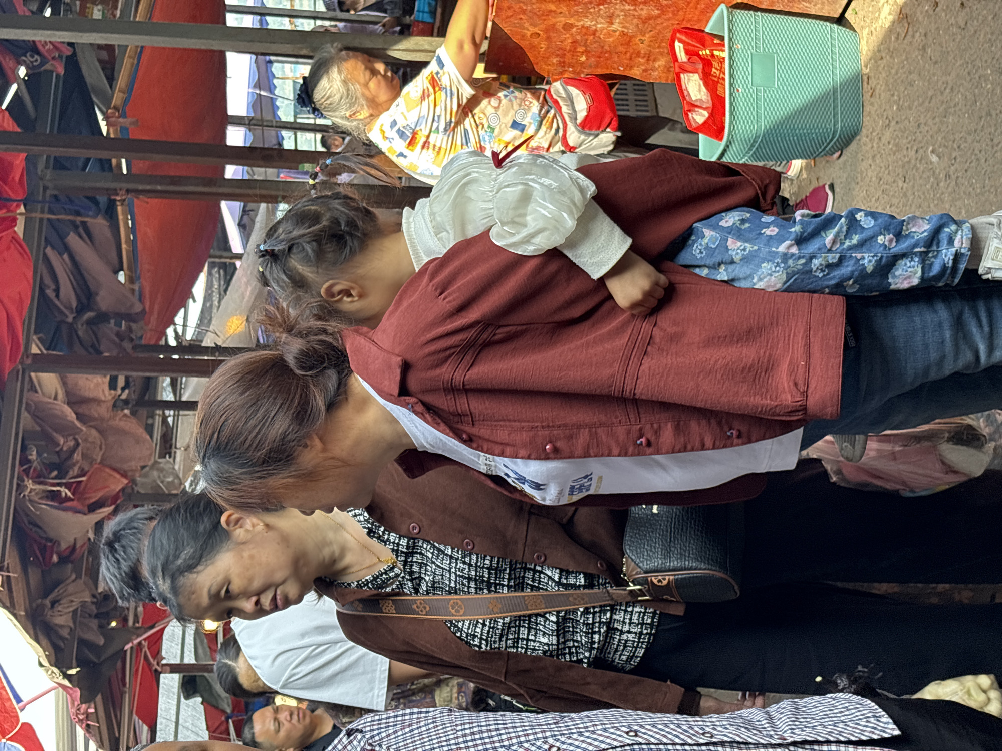 The image depicts a street scene in Jiangjin, China. Three generations of women are visible: an elderly woman in a patterned shirt sitting at a table with a green basket; a younger woman wearing a brown jacket over a checkered shirt and a shoulder bag; and a child in a white blouse and floral pants being carried. The background includes various market stalls with red and blue awnings, and assorted goods for sale.