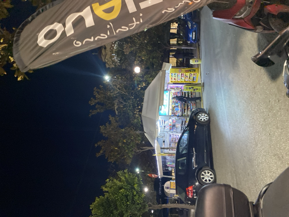 This nighttime outdoor scene, captured in Egaleo, Greece, depicts a street with various activities under artificial illumination. The image is oriented sideways, but when viewed upright, it reveals a bustling area.
In the upper left, a large, dark fabric banner hangs, partially obscuring the dark night sky. It displays white and yellow text; the white letters clearly show "ON" with mirrored Greek script "ανιλτι" (anilti) below, suggesting a brand name or slogan.
The central focus is a brightly lit street stall or kiosk, covered by a white tent canopy. The interior of the stall is visible, showcasing shelves packed with an array of merchandise including snacks, drinks, and various small items. A person is discernible inside the stall, possibly attending to customers or arranging products.
Adjacent to the stall, a dark-colored hatchback car is parked. Further into the background, a blue car is partially visible, and on the right side of the frame, parts of a red and a black scooter or motorcycle are also parked.
The scene is framed by trees, with their foliage visible in the background and extending into the lower left corner. Streetlights dot the area, casting light and creating visible flares against the dark sky. A smaller, rectangular sign is partially visible in the lower-left background, displaying the word "CERTIFUO". The overall atmosphere suggests a lively commercial street operating into the evening.