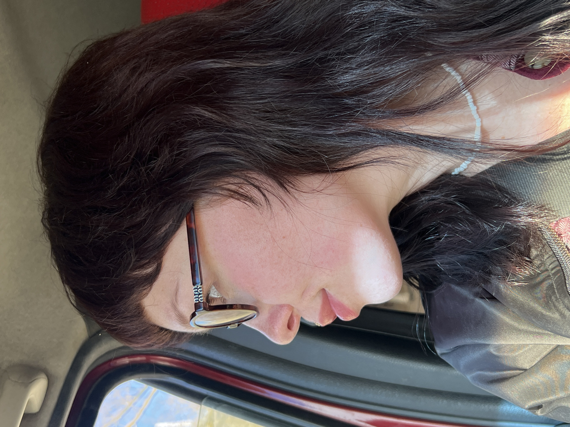 A close-up, side profile of a person, likely female, is depicted, oriented horizontally within a vehicle. The individual has fair skin, adorned with light freckles and a natural rosy blush on the cheek. They wear tortoise-shell framed glasses, featuring a metallic, patterned detail on the temple. Their long, dark brown hair, appearing thick and slightly wavy with some lighter strands, flows around their face and neck. A delicate, light-colored (possibly white or translucent) beaded necklace or string is visible around the neck, partially covered by hair. The person's lips are slightly parted, and their expression conveys a sense of calm and neutrality.
The setting appears to be the interior of a car, indicated by the reddish-maroon trim of a car door frame and a window reflecting a bright, sunny sky with indistinct shapes. Part of a dark grey or olive-colored garment, possibly a jacket with a zipper, is visible on the right side of the frame. The strong, natural lighting suggests it is daytime. The person seems to be resting or traveling as a passenger. No specific activity or event is explicitly taking place. There is no clearly legible text visible on any objects within the frame. The location context provided is Izhevsk, Russia.