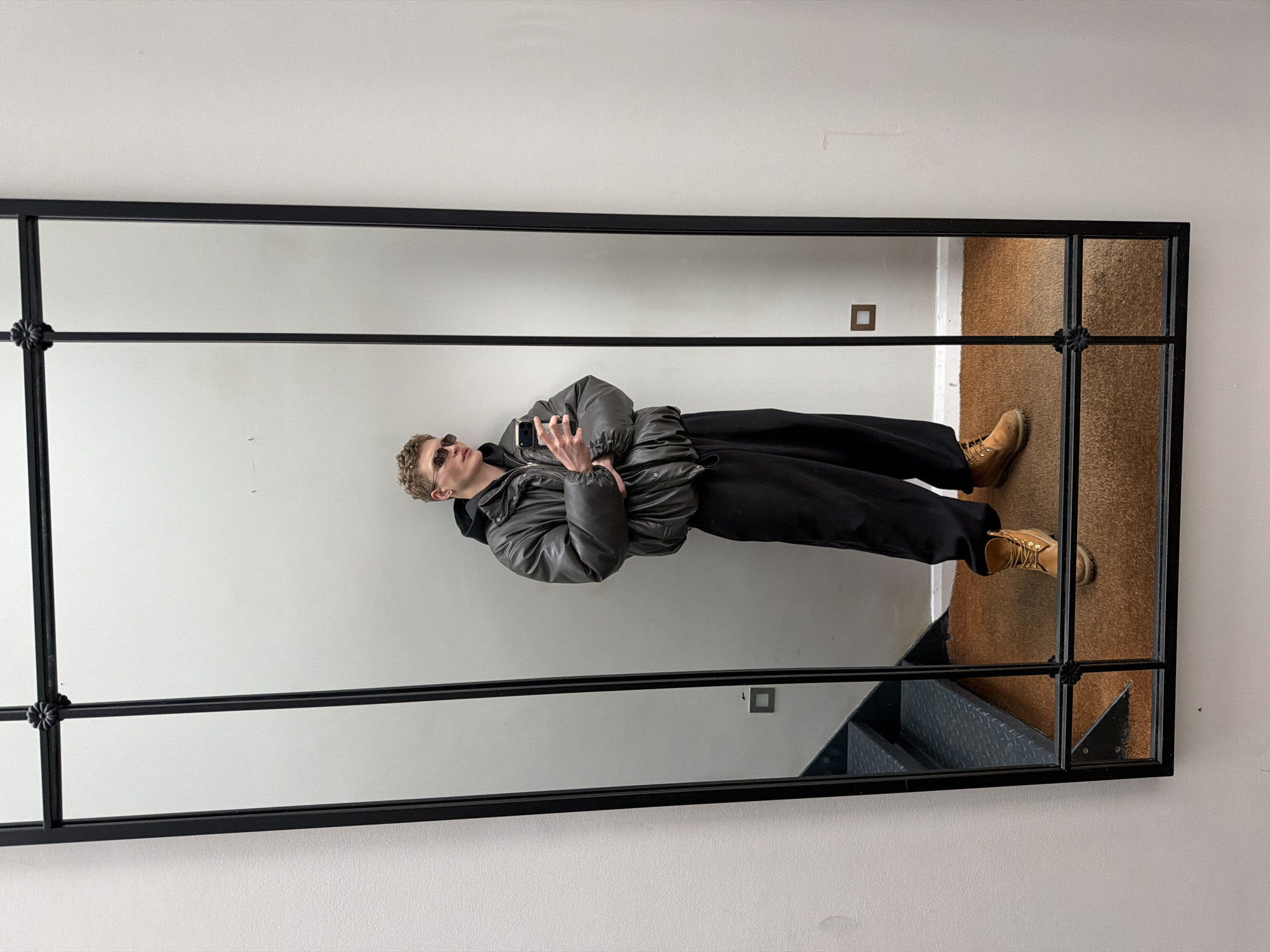 A mirror reflection of a man in a grey puffer jacket, black pants, and tan boots. He is wearing sunglasses and holding a phone. He is standing in front of stairs in what appears to be an indoor location. It is not possible to determine if the location is in London, United Kingdom from the image alone.