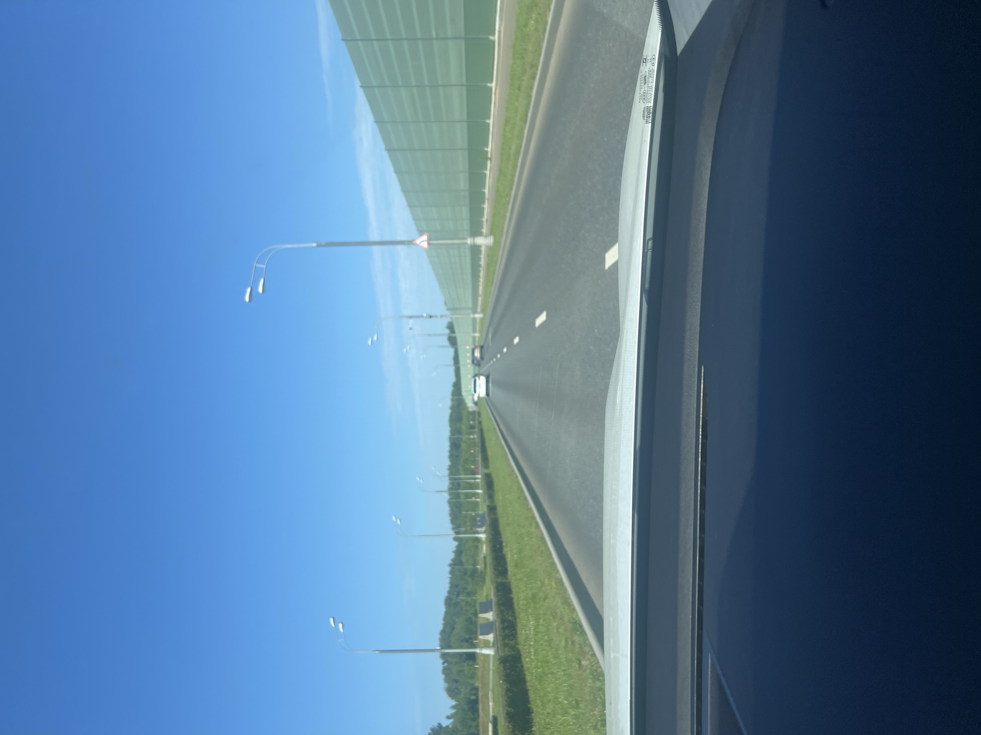The image shows a view from inside a vehicle traveling on a highway, likely in or near Cherepovets, Russia. The road is asphalt with white lane markings. On the right side of the road, there is a tall, pale green sound barrier. On the left side of the road, there is a grassy embankment lined with hedges, followed by trees and a blue sky. Several streetlights are visible along the roadside, and a few cars are in front of the vehicle, moving in the same direction. The dashboard of the vehicle is visible in the foreground, reflecting the sky and road.