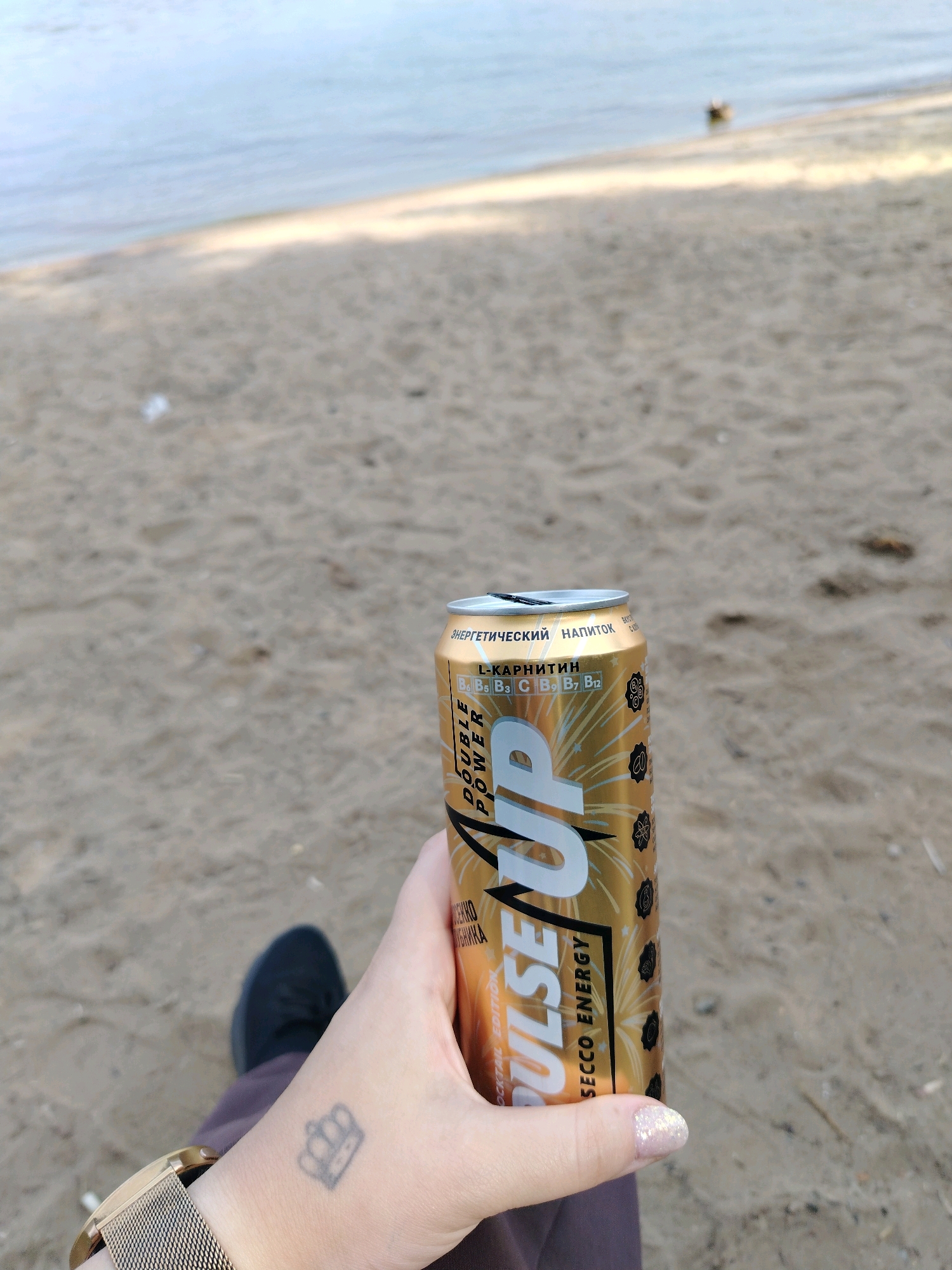 The image shows a person's hand holding a can of "UP DOUBLE POWER" energy drink on a sandy beach next to a body of water. The can is gold with white lettering and a design of fireworks. The person's wrist has a small tattoo of a crown and they are wearing a metallic watch. In the background, the water appears calm and the sand is light brown. The overall scene suggests a relaxed outdoor setting.