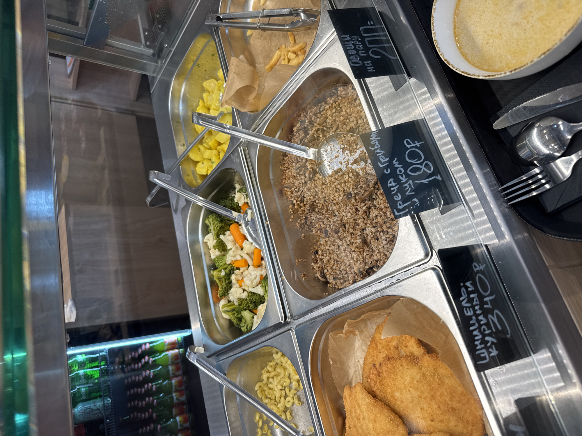 The image shows a cafeteria-style food display, likely in Yakkima, Russia. In the foreground, a white bowl of creamy soup is visible, with a fork, spoon, and knife set to its left. Behind the bowl, a metal tray holds a portion of cooked buckwheat with mushrooms, and a handwritten sign indicates "Греча с грибами и луком 180р" (Buckwheat with mushrooms and onions 180 rubles). To the right of the buckwheat is another metal tray with french fries and a pair of tongs. A third sign above the french fries reads "Овощи на пару 200" (Steamed vegetables 200). Further back and to the left, a tray contains breaded chicken cutlets with a sign reading "Шницель куриный 340р" (Chicken schnitzel 340 rubles). Above the cutlets, a tray holds boiled pasta. Another tray to the right contains a mixture of steamed broccoli, cauliflower, and carrots. In the background, a refrigerated display case is filled with green bottles of soda, likely Sprite, and other beverages. The overall environment appears to be a clean, well-lit food service area. No people are visible in the image.