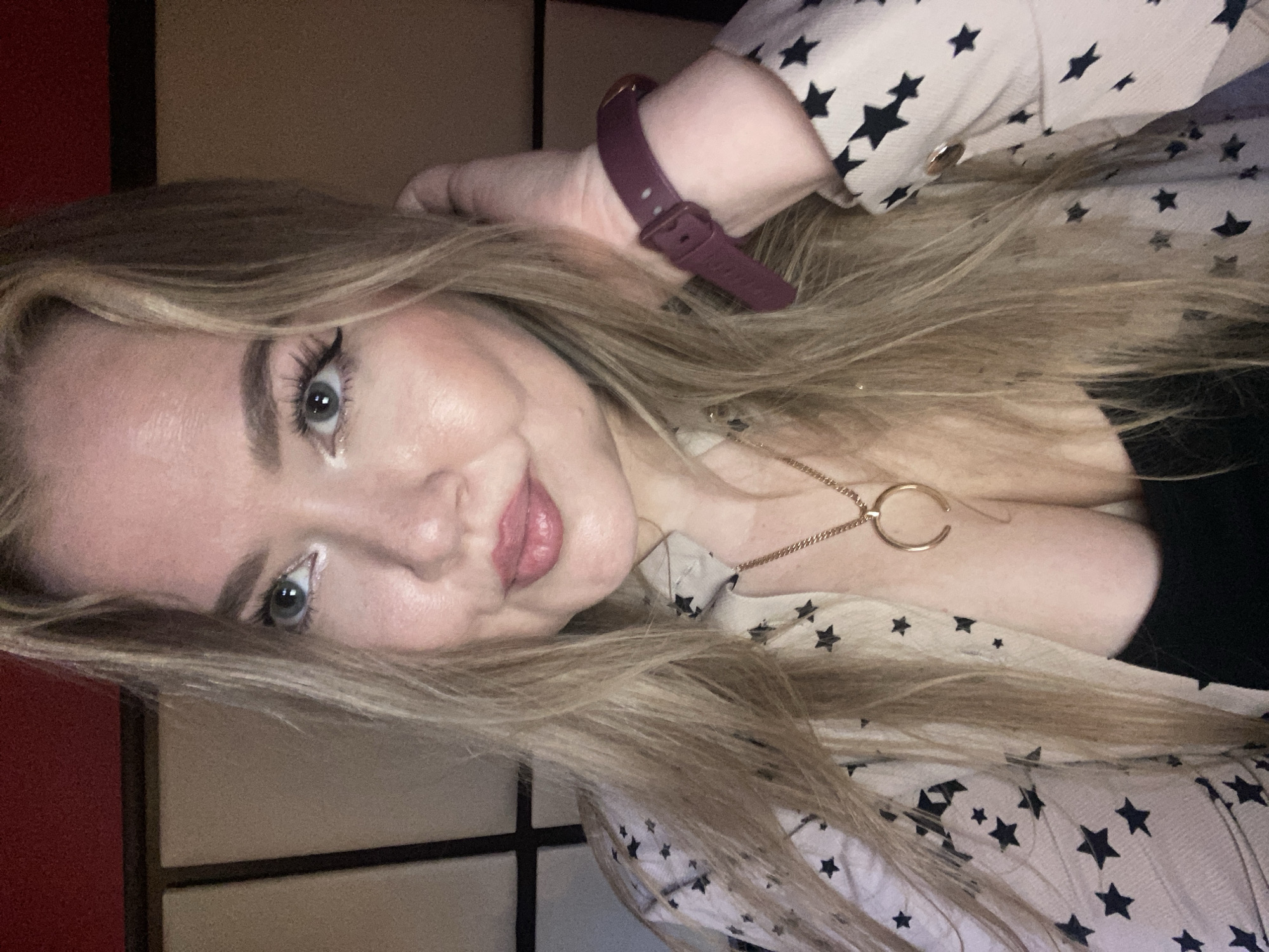The media file shows a close-up, sideways portrait of a young woman with long, blonde hair. She is wearing a light-colored shirt with a pattern of black stars and a maroon-colored watch on her left wrist. A delicate gold necklace with a circular pendant is visible around her neck. Her makeup includes eyeliner and mascara, and her lips are painted a pinkish hue.
The lighting suggests an indoor setting, possibly in the evening or during the day with artificial lighting. The background appears to be a room with panels, with a reddish element on the left and a neutral-toned panel on the right. There are no visible signs of weather or time of day beyond the general indoor lighting. The woman's expression is relaxed and she appears to be looking towards the camera. The location context is Yerevan, Armenia, but there are no specific visual cues within the image to confirm this.
