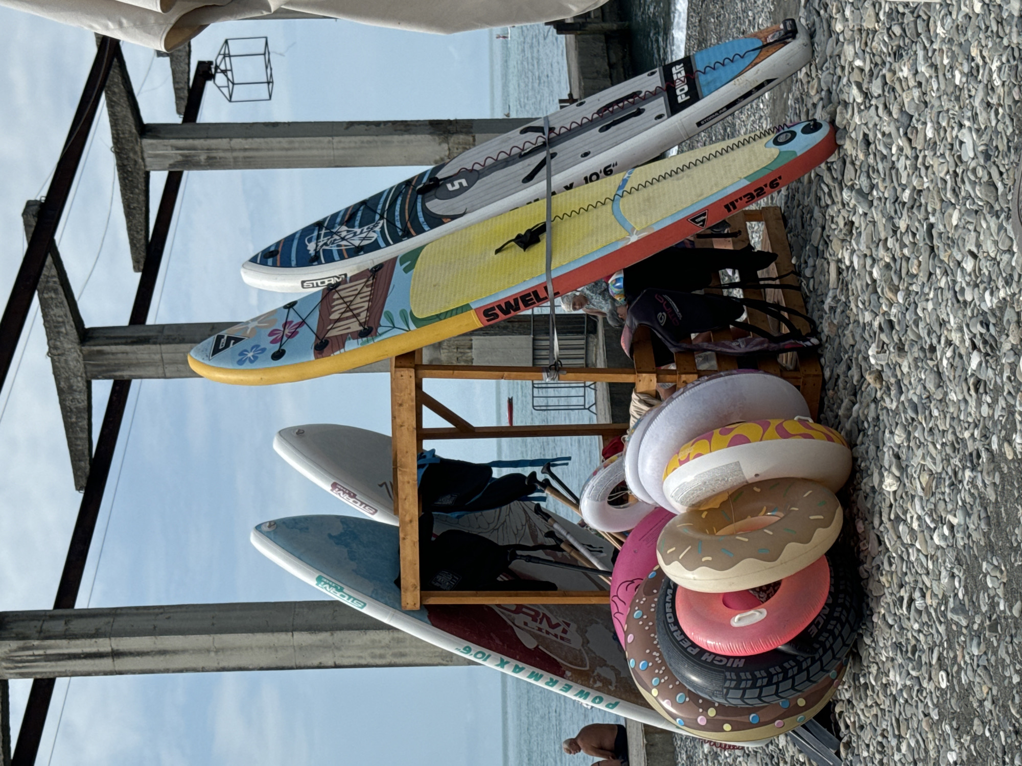 The image shows a collection of stand-up paddleboards and inflatable rings stacked on a wooden rack by a pebble beach. The paddleboards are of various colors and designs, with some bearing brand names like "STORM" and "POWERMAX." The inflatable rings are shaped like donuts and are stacked on top of each other. In the background, the sea is visible, suggesting a beachside location. The overall scene depicts a recreational area, likely at a beach resort. The lighting suggests it is daytime, possibly late afternoon or early evening due to the shadows. There are no people prominently featured, though a man is partially visible in the bottom right corner. The weather appears to be clear and sunny. The text visible on the paddleboards includes "STORM," "POWERMAX 10'6"," "POWER," and "SWEL." The inflatable rings have text that is partially visible, including "HIGH PERFORMANCE." The setting appears to be Sochi, Russia, given the context provided.