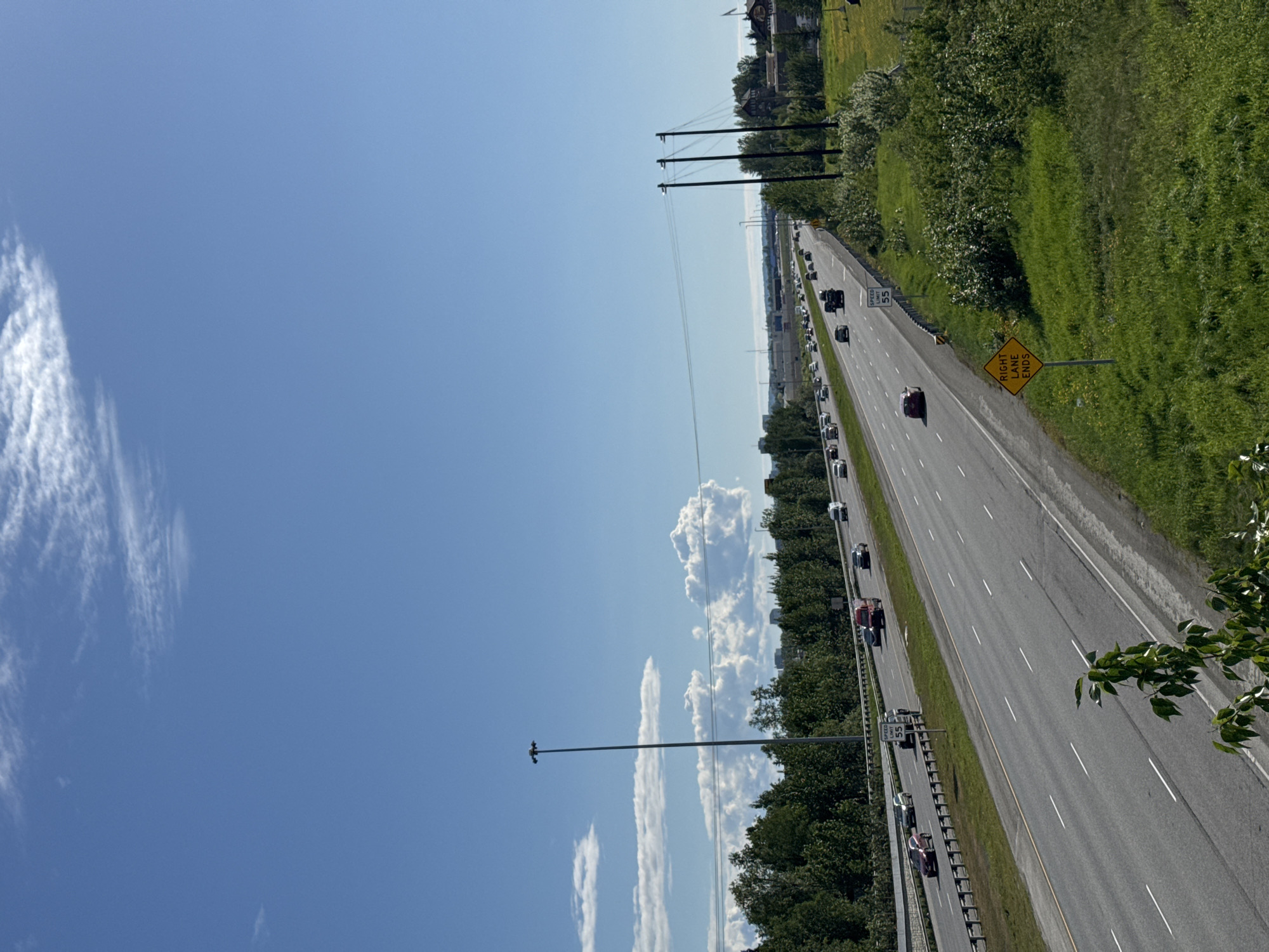 This is a photograph taken from an elevated vantage point, looking down upon a multi-lane highway in Anchorage, Alaska, United States. The sky is a vibrant blue with scattered white clouds. The highway, appearing to be a section of the Seward Highway, is filled with a moderate amount of traffic. Several vehicles, including cars and a red truck, are visible on the roadway. On the right side of the highway, a green grassy embankment is present, with scattered bushes and trees. A yellow diamond-shaped sign indicating "RIGHT LANE ENDS" is visible in the foreground, along with a speed limit sign for 55 mph. On the left side of the highway, there are more trees and some buildings in the distance. Power lines and a tall street light are also visible. The overall impression is a typical highway scene in a suburban or semi-urban environment on a clear day.