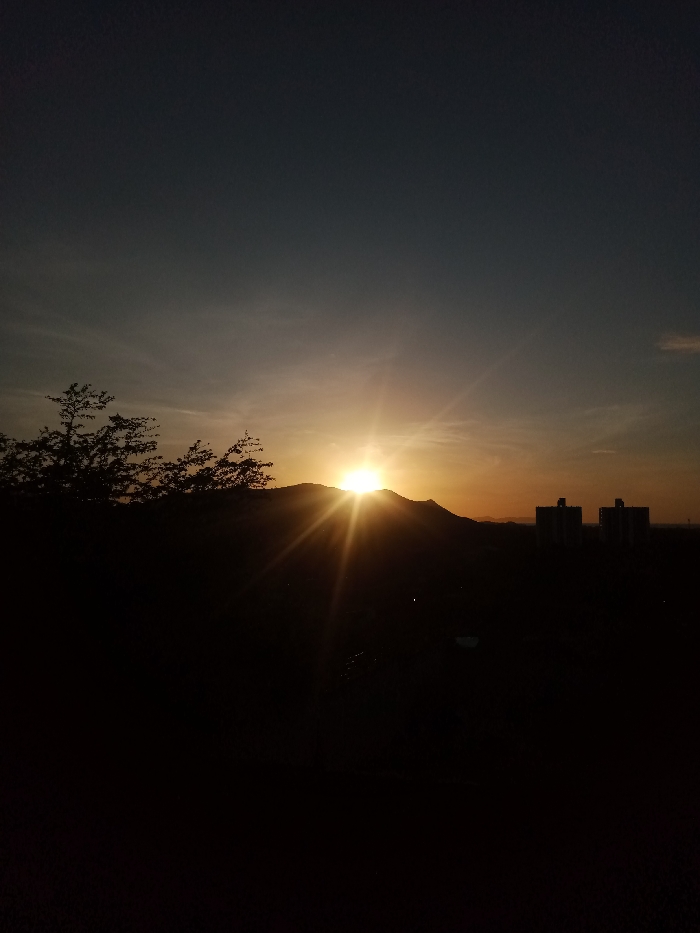 A vibrant sunset illuminates the horizon, its intense glow peeking over a silhouetted mountain range. The sky transitions from a deep blue at the top to a warm orange near the horizon. A dark tree line is on the left, while two indistinct buildings stand on the right. The foreground is shrouded in deep shadow, adding depth and contrast to the sun-drenched scene.