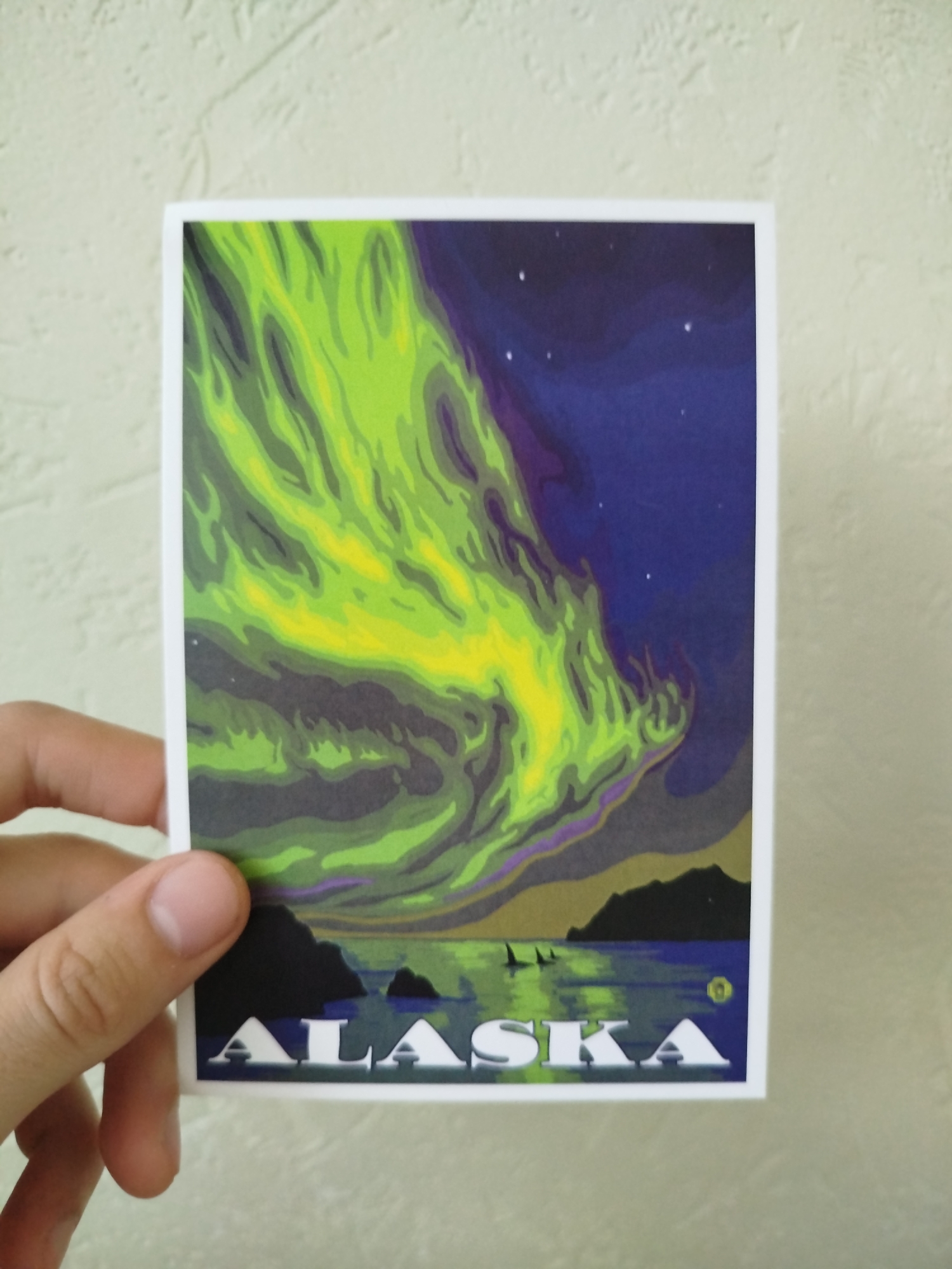 A hand holds a postcard with an illustration of the aurora borealis over a body of water with land in the background. The word "ALASKA" is printed at the bottom of the postcard. The background is a light beige wall. The location is unknown, but there is no indication that the image is in Varna, Bulgaria.