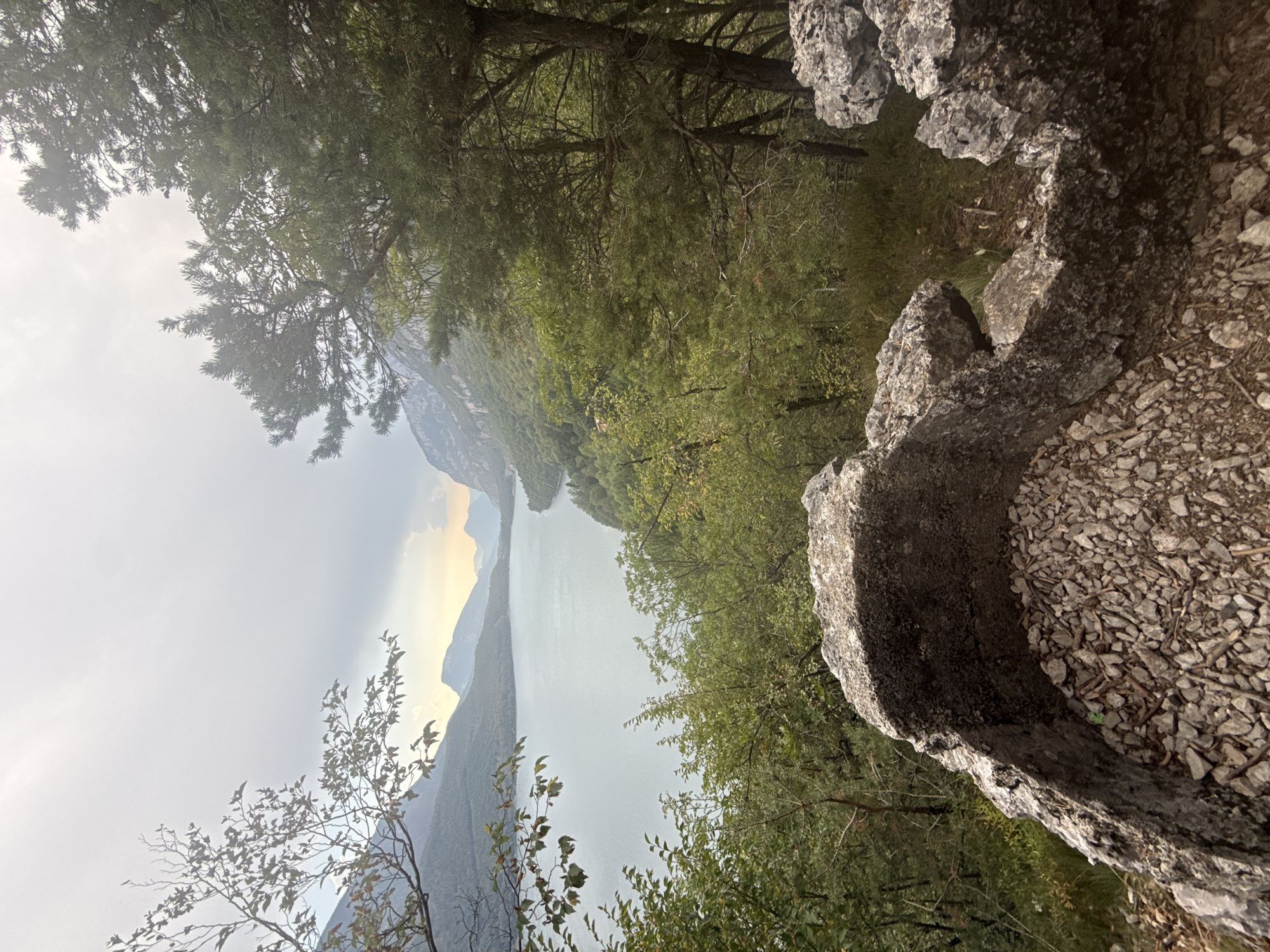 An outdoor, high-angle, medium shot captures a serene mountain lake vista framed by trees and rocks. The lake, calm and vast, reflects the overcast sky. Jagged, grey rocks dominate the foreground, their textured surfaces filled with smaller stones and debris, suggesting a natural, perhaps eroded, formation. Dense, green foliage from trees and bushes flanks the lake and fills the midground, with some branches and leaves reaching into the frame from the left and right edges. In the distance, the lake narrows and curves, with imposing, misty mountains rising on either side, their peaks softened by atmospheric haze. The sky above is a pale, diffused grey, with subtle hints of lighter tones suggesting a muted sunrise or sunset. The scene is devoid of any human presence.