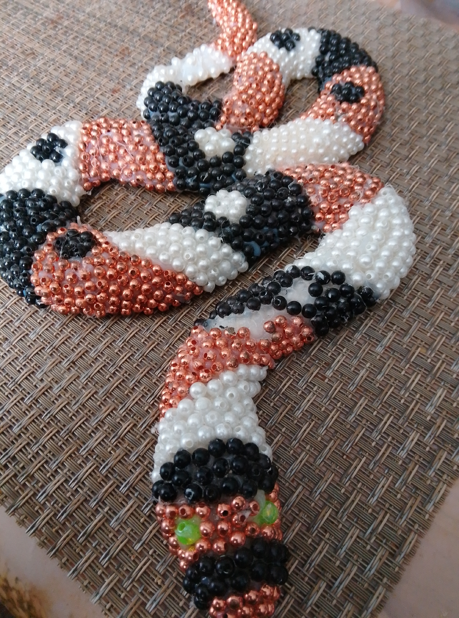 A close-up, overhead shot captures a beaded snake crafted from white, black, and copper-colored beads. The snake is intricately coiled, with its head facing the bottom right corner of the frame. Two small, bright green beads serve as eyes, and a few black beads form its snout. The snake rests on a textured, brown placemat, likely in a home setting. There are no people or any identifiable landmarks visible. This image provides no information about the city of Wedel, Germany.