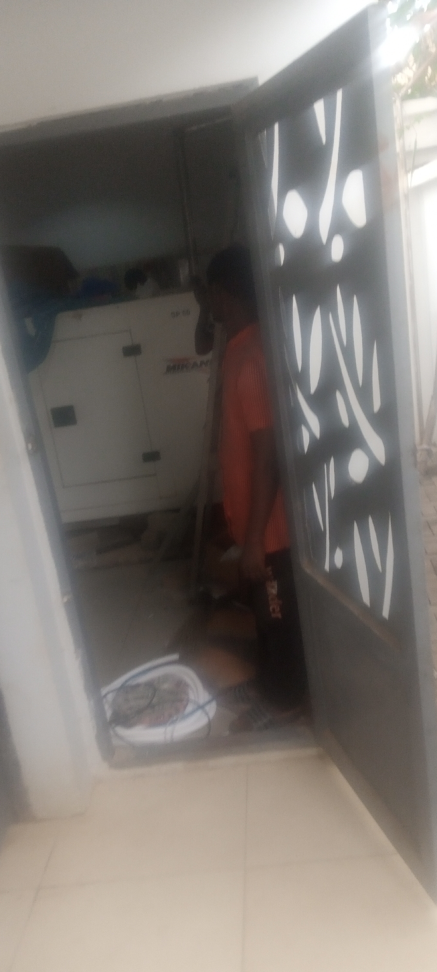 The image shows a man standing inside a generator room, accessible through an ornate metal gate. Inside the room is a large white generator labeled "MIKANO". The man is wearing an orange shirt and black pants with a visible "Adidas" logo. A white plastic bowl containing coiled material (possibly electrical wiring) is on the floor near the entrance. The room appears to be part of a building with tiled floors. Based on the prompt, the location is Unguwar Rimi, Nigeria.